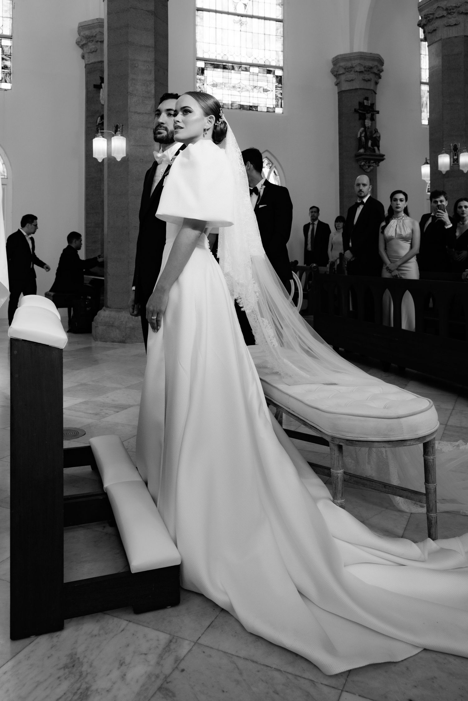 Black and white photo of a bride in elegant gown with cape veil at church altar during wedding ceremony.