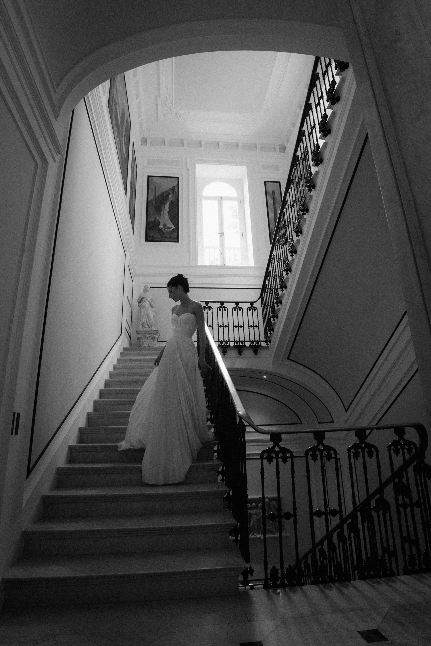 Elegant bride in a flowing white gown descends a grand staircase in a classic mansion in black and white.