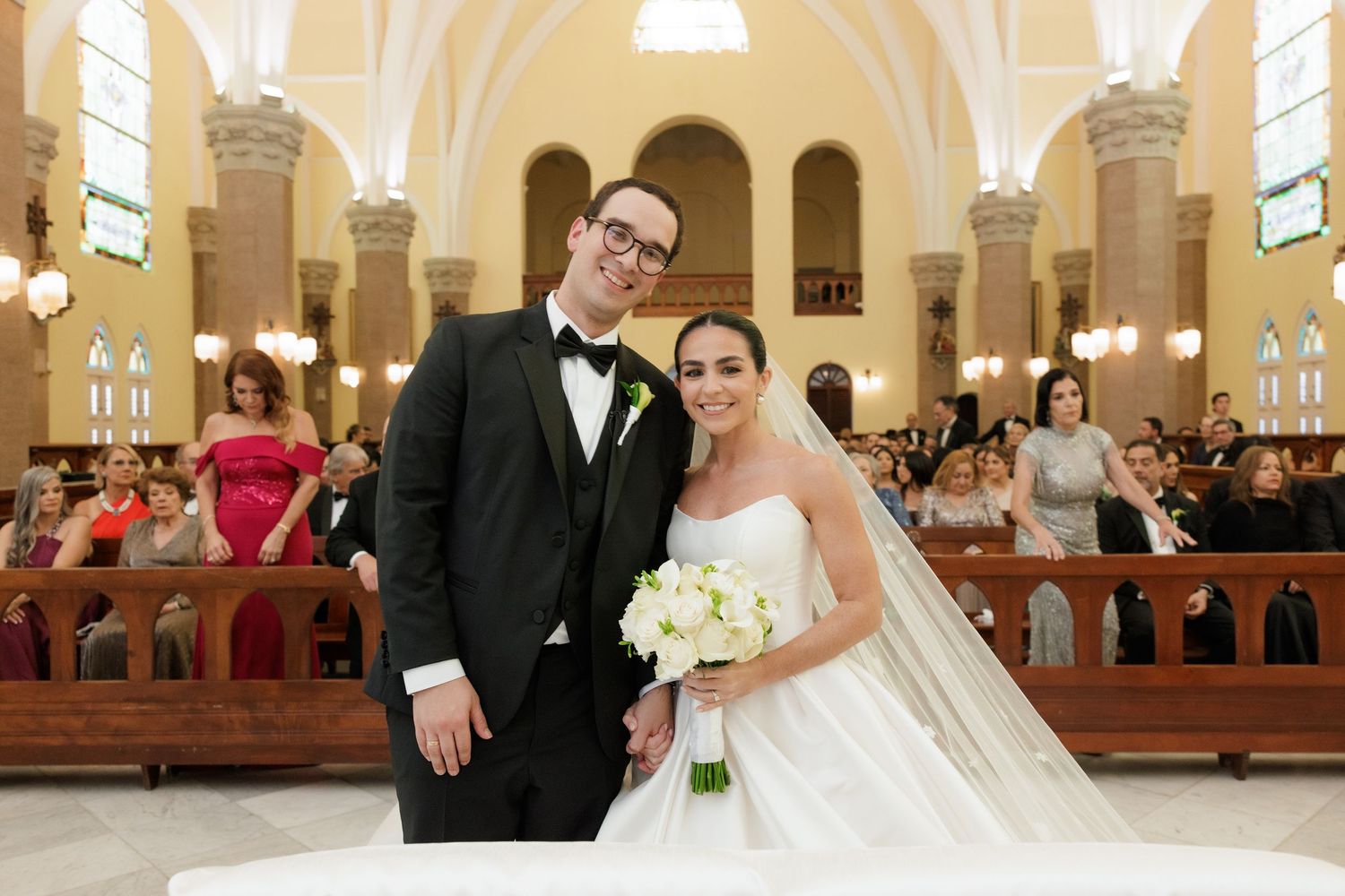 A wedding couple poses in front of church pews with ornate architectural details visible in background.