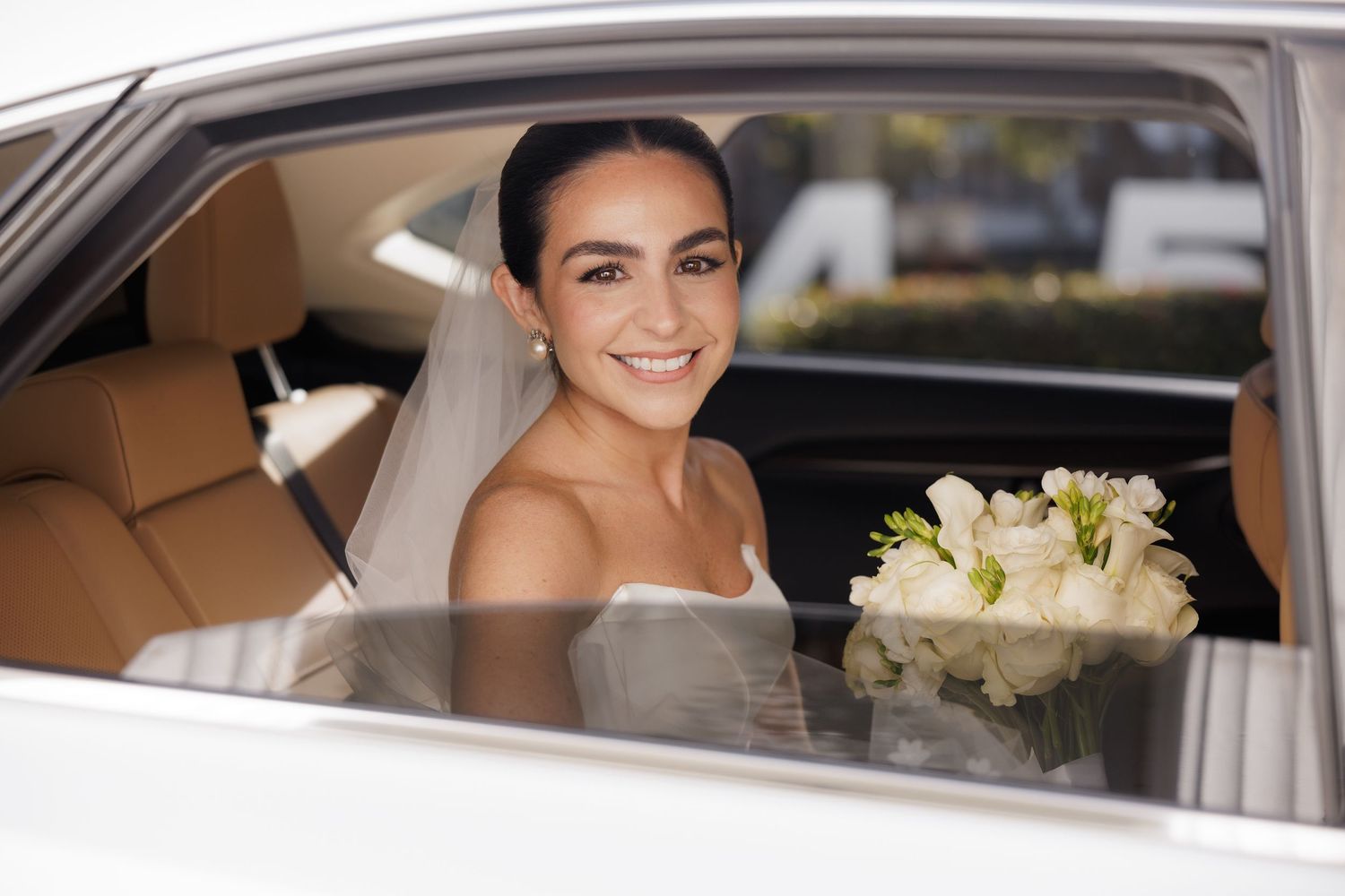 A bride in a strapless wedding dress sits in a luxury car with a white bouquet.