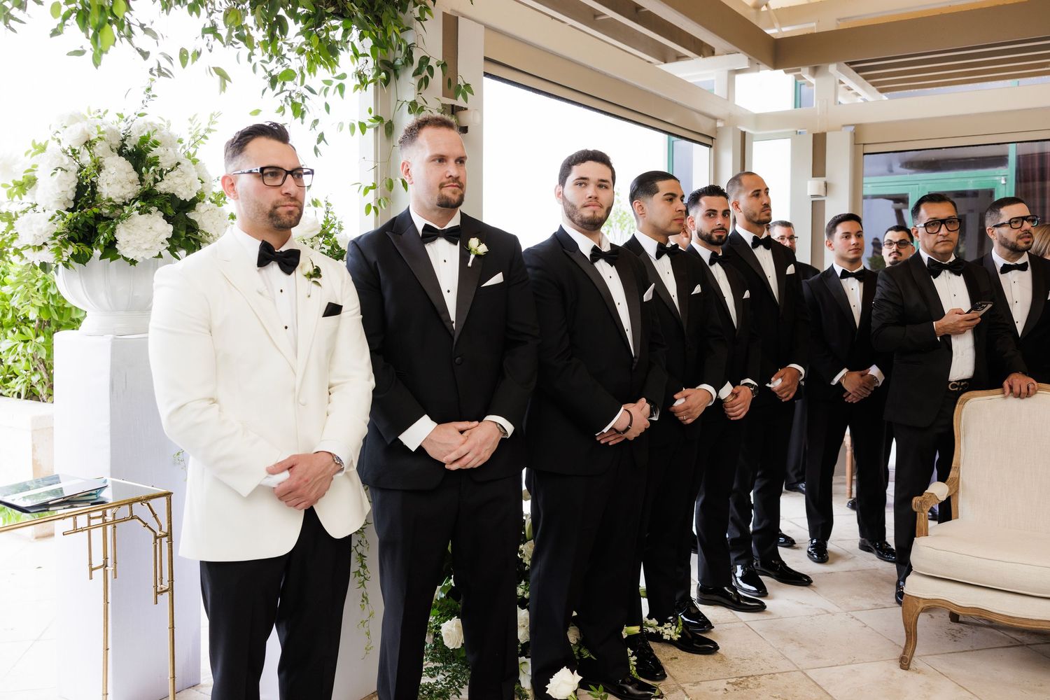 Wedding party poses outdoors with groomsmen in black tuxedos and bridesmaids in dark dresses.