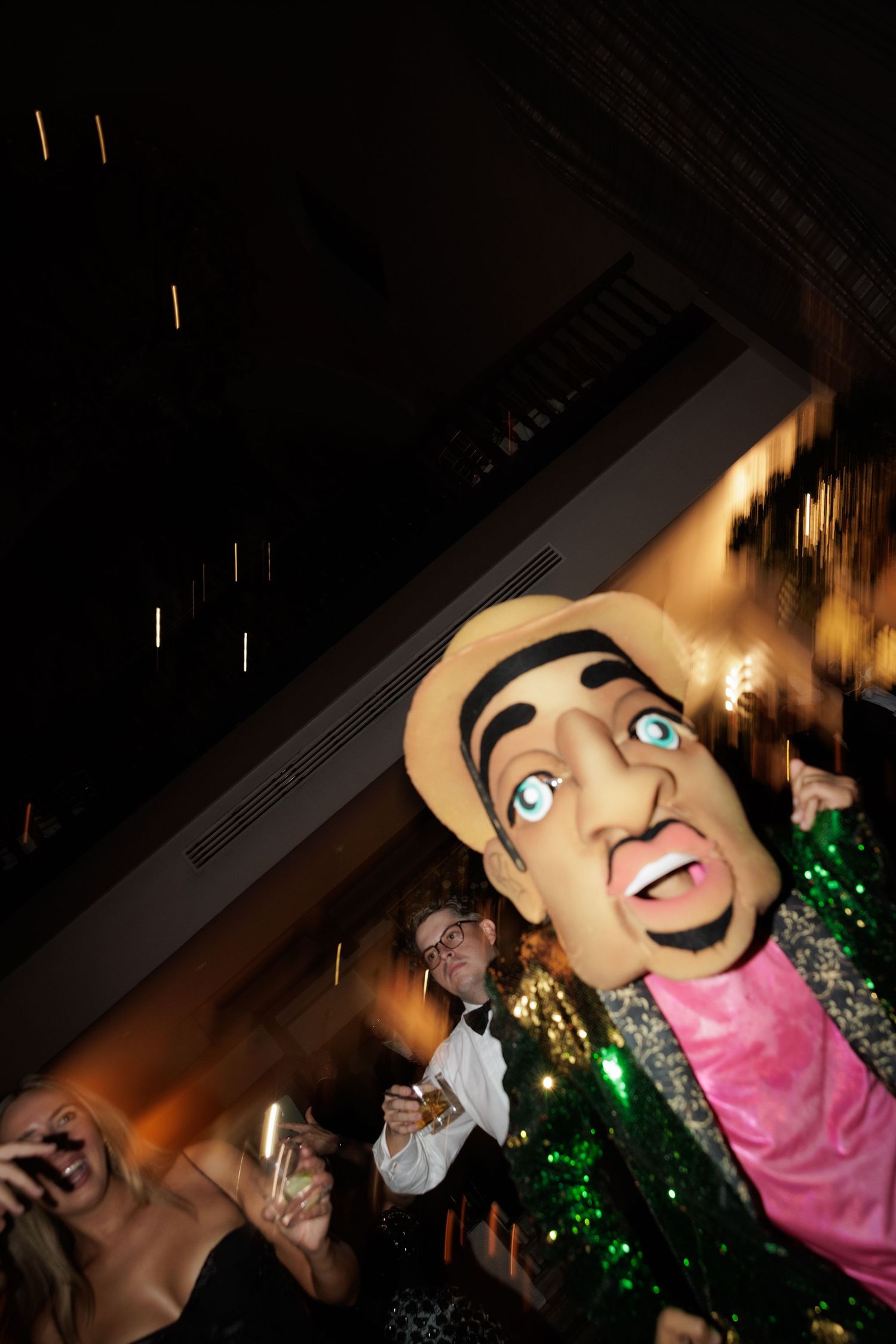 Large puppet-like mascot heads with exaggerated features appear in a dimly lit party setting.