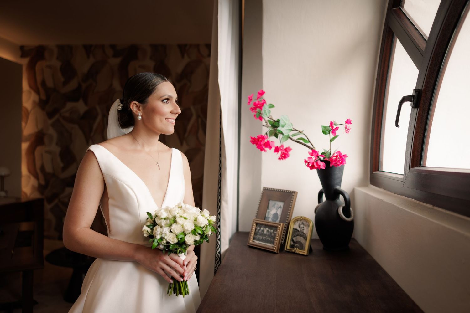 Bride in elegant white V-neck dress holds bouquet while standing near window with pink flowers in vase.