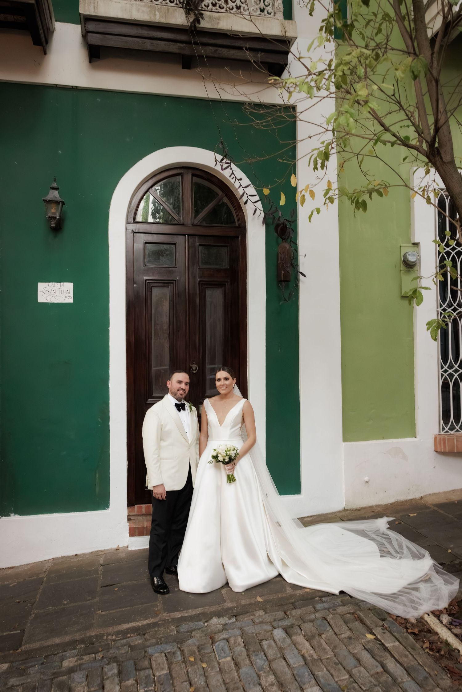 A wedding couple poses against green and white colonial walls and ornate doorways on a cobblestone street.