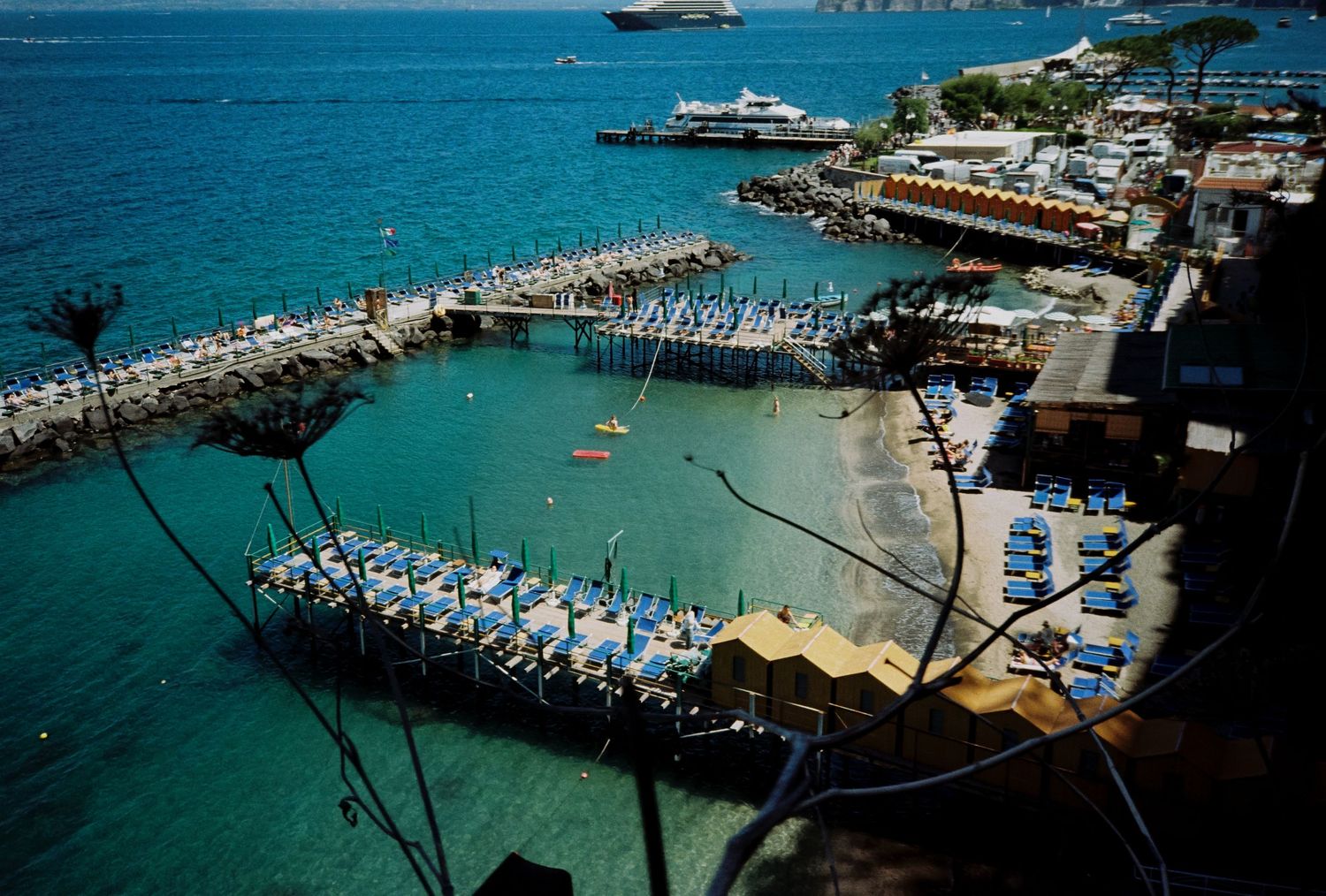 Aerial view of a Mediterranean harbor with blue lounge chairs, boats, and turquoise water near a rocky pier.