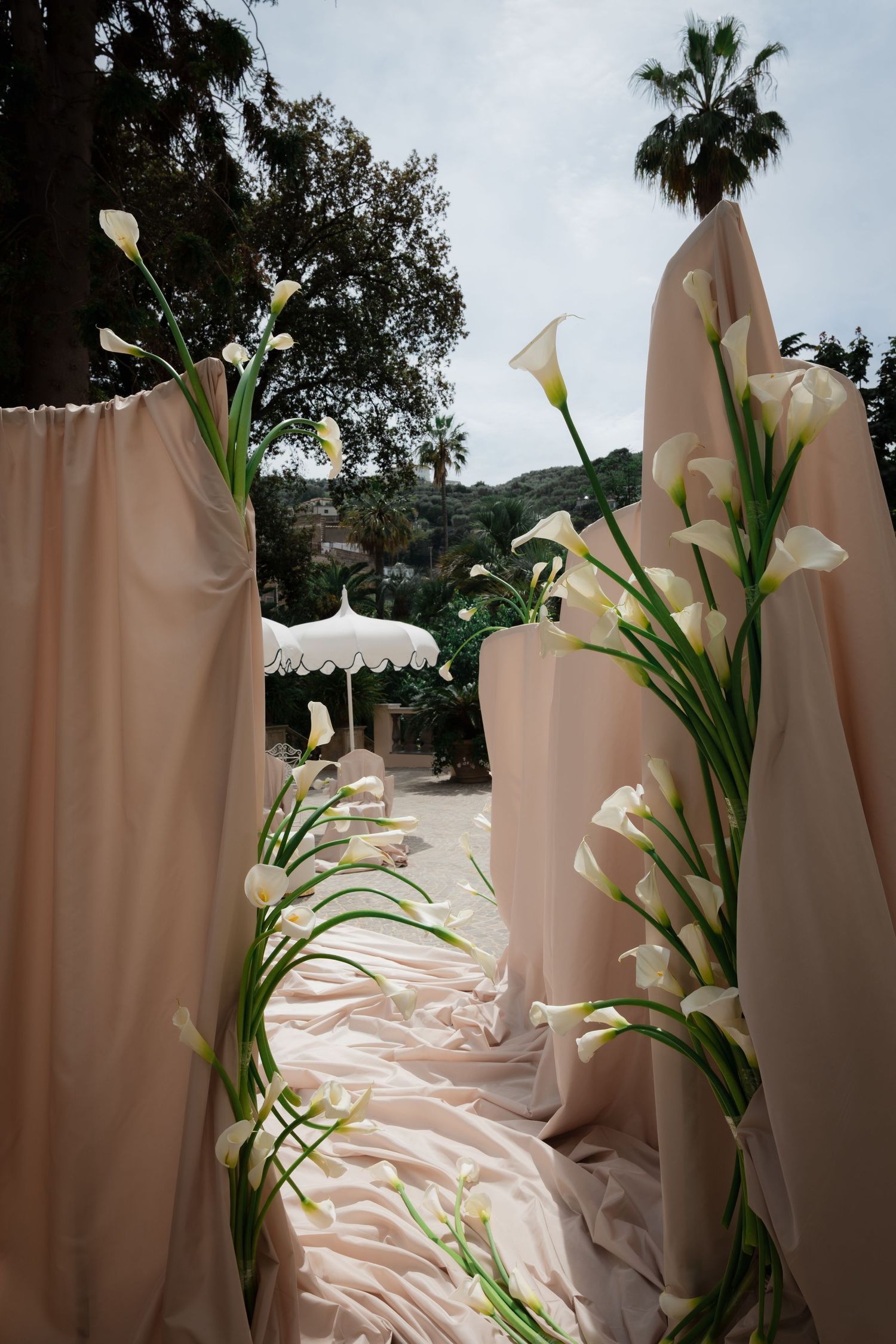 Elegant outdoor wedding aisle with blush draping and white calla lilies lining the path, surrounded by lush greenery.