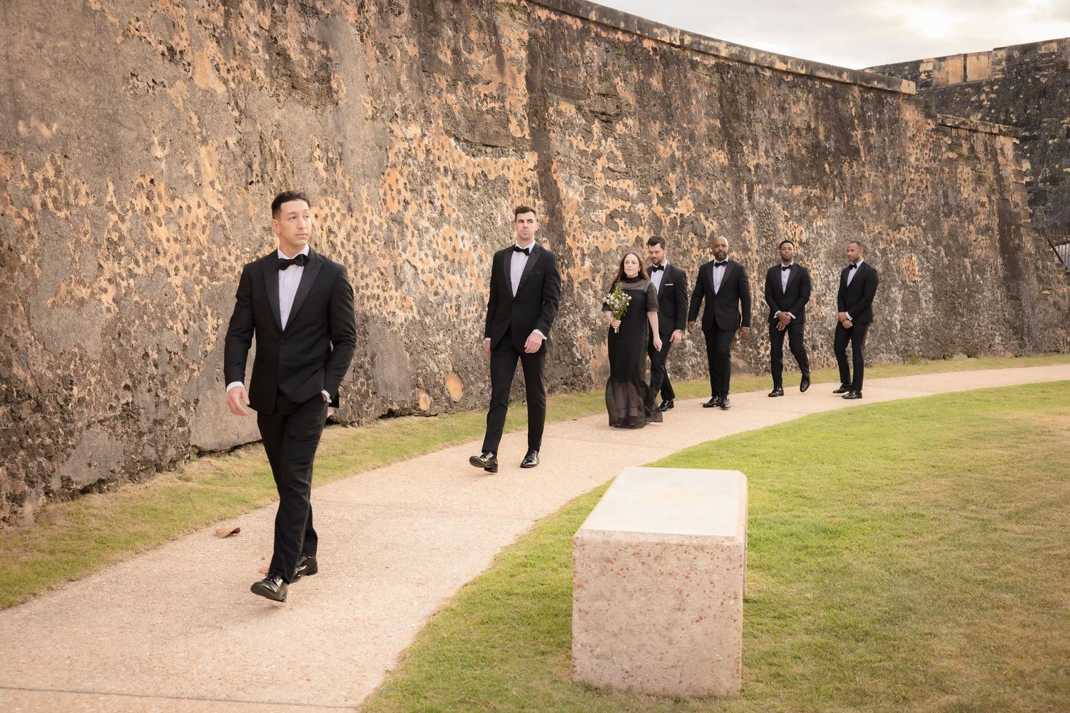 Groomsmen in black tuxedos walking along stone fortress wall during wedding photography session.