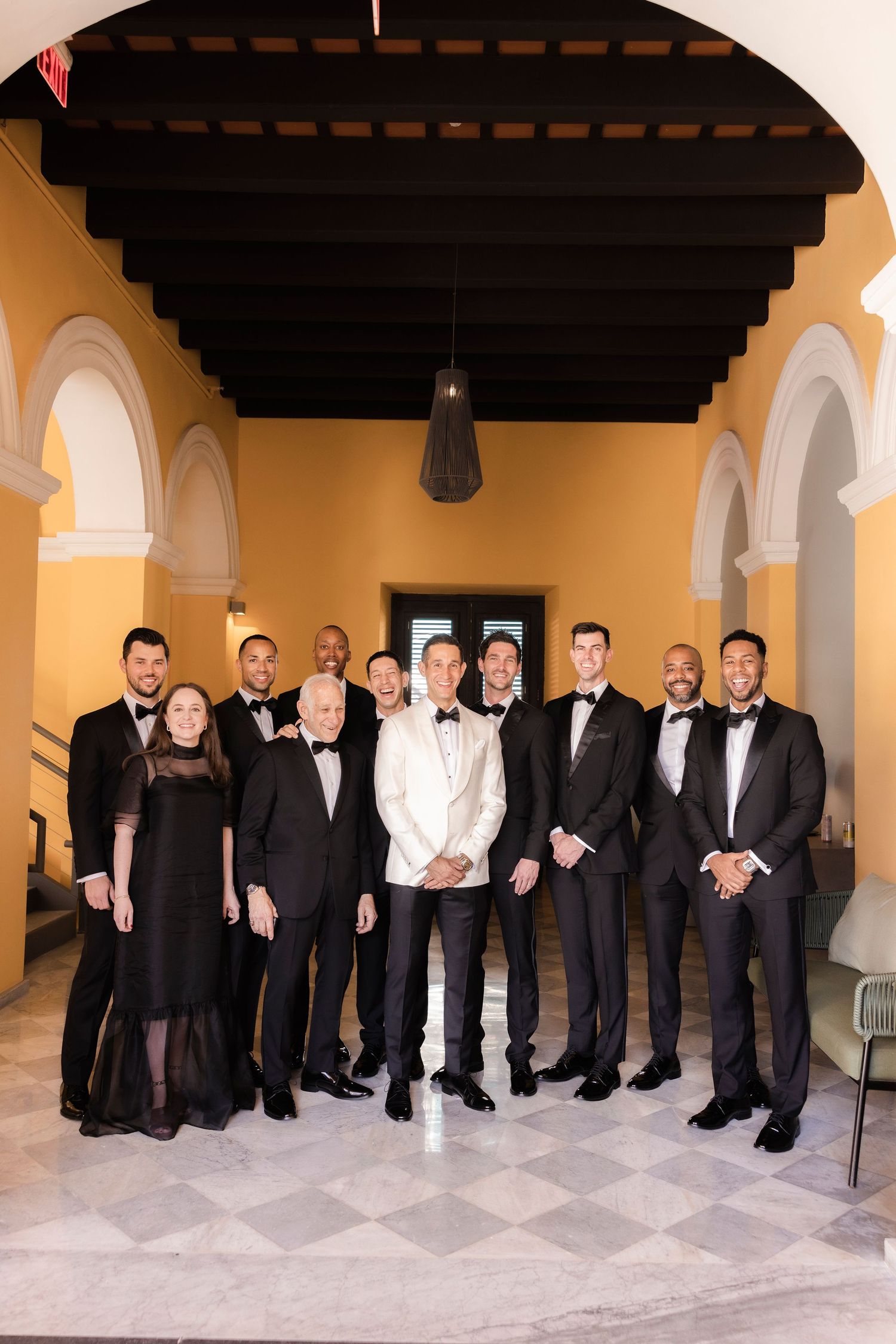 Large wedding party in formal black and white attire posing in an arched hallway.