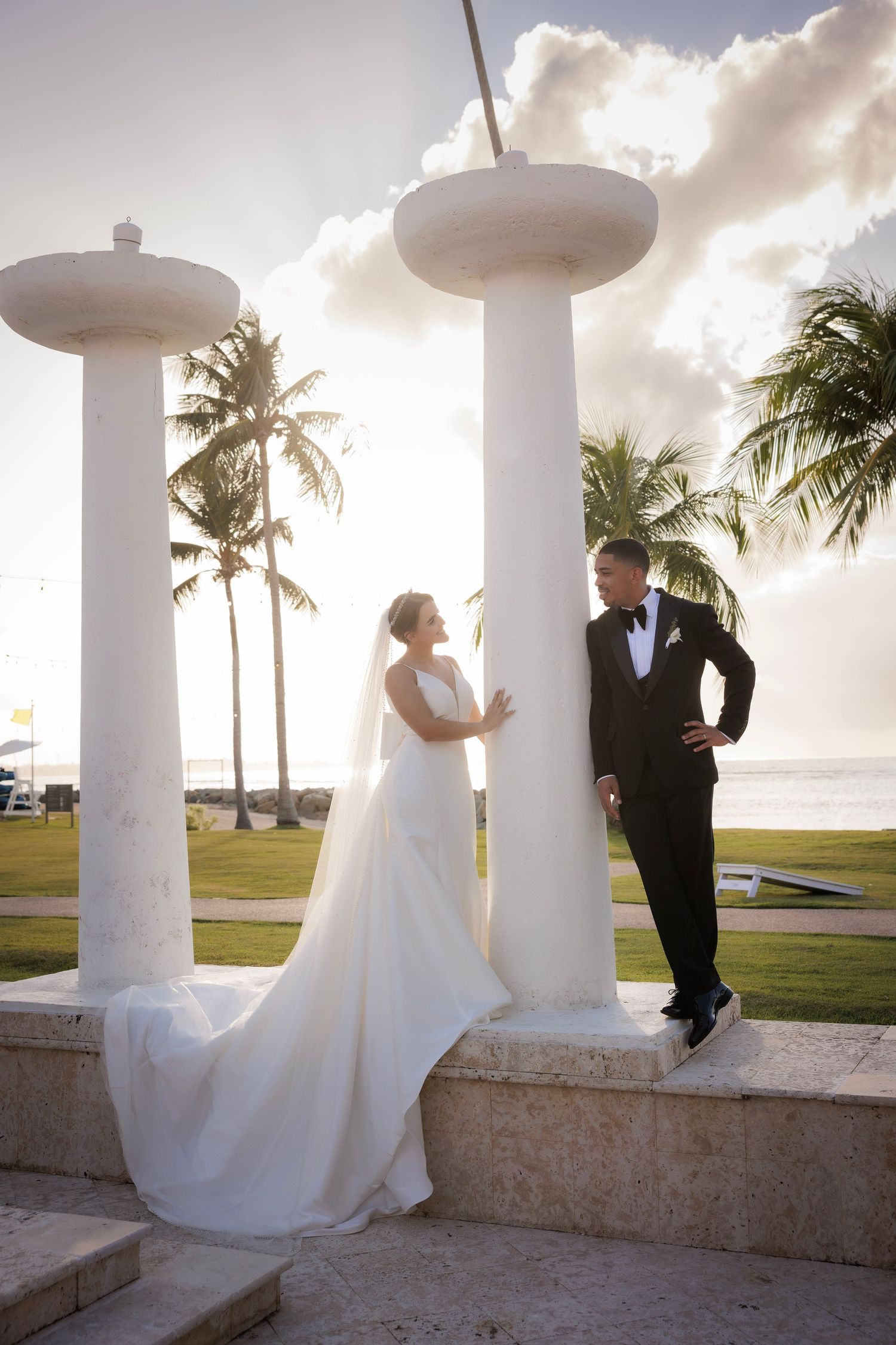 A romantic wedding couple shares a moment between white columns at sunset with palm trees in the background.