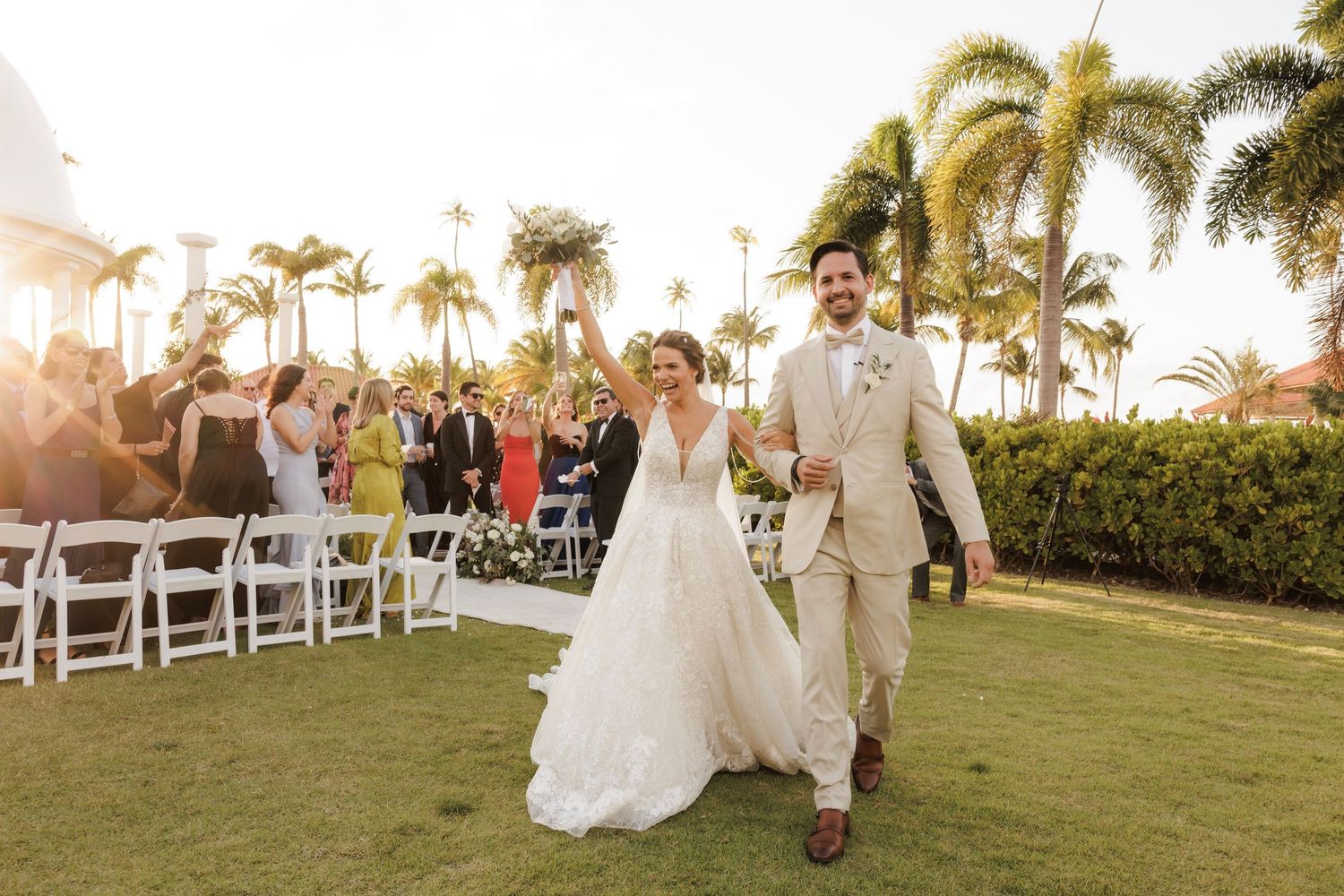 Sun sets behind an elegant outdoor wedding celebration with palm trees and white folding chairs on a lush lawn.
