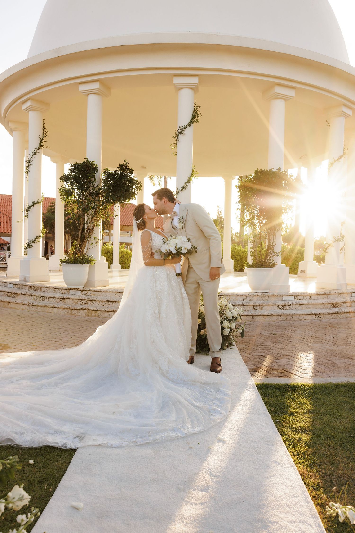 Long flowing white wedding dress trail extends across a garden patio with white columns at sunset.