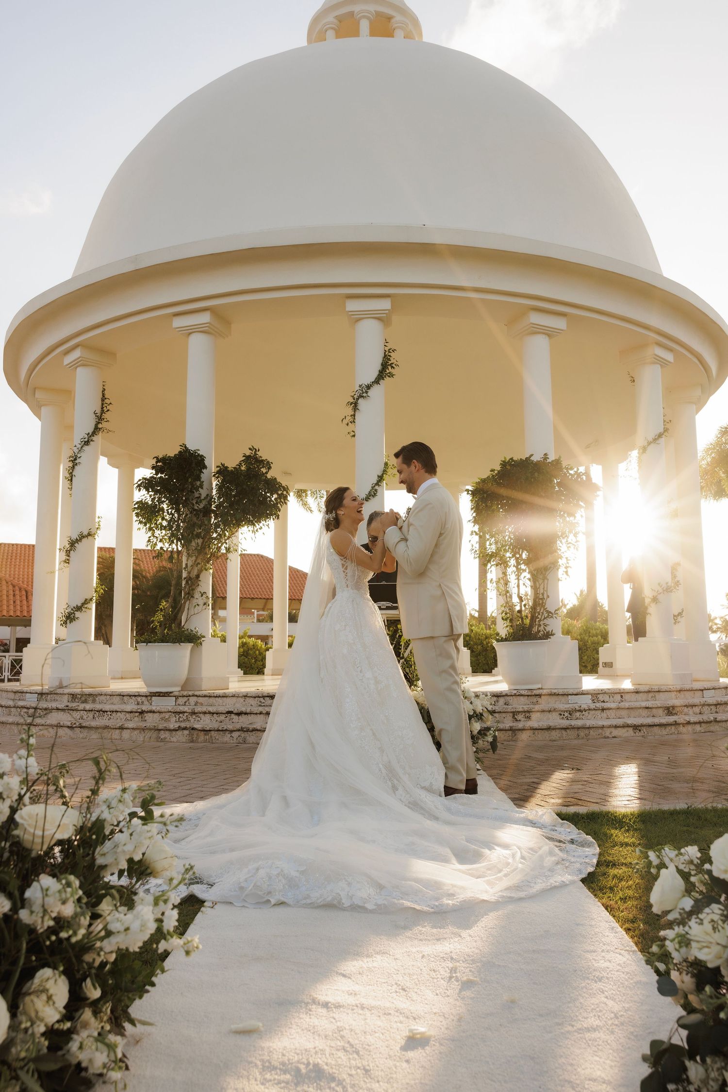 Elegant bride in flowing white gown poses beneath a romantic circular gazebo at sunset with garden landscaping.