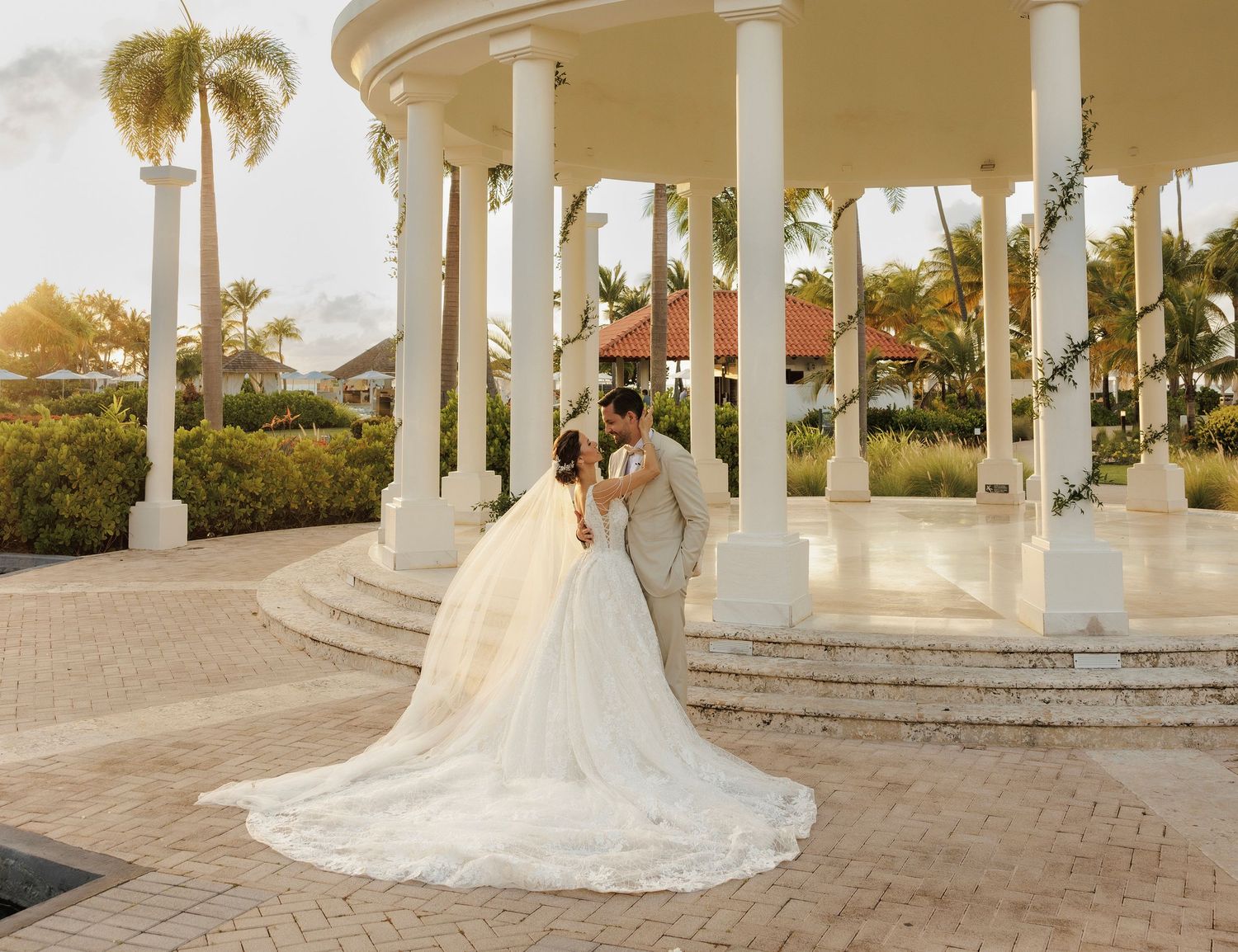 A happy couple poses in front of an elegant white pavilion with palm trees during their outdoor wedding photoshoot.