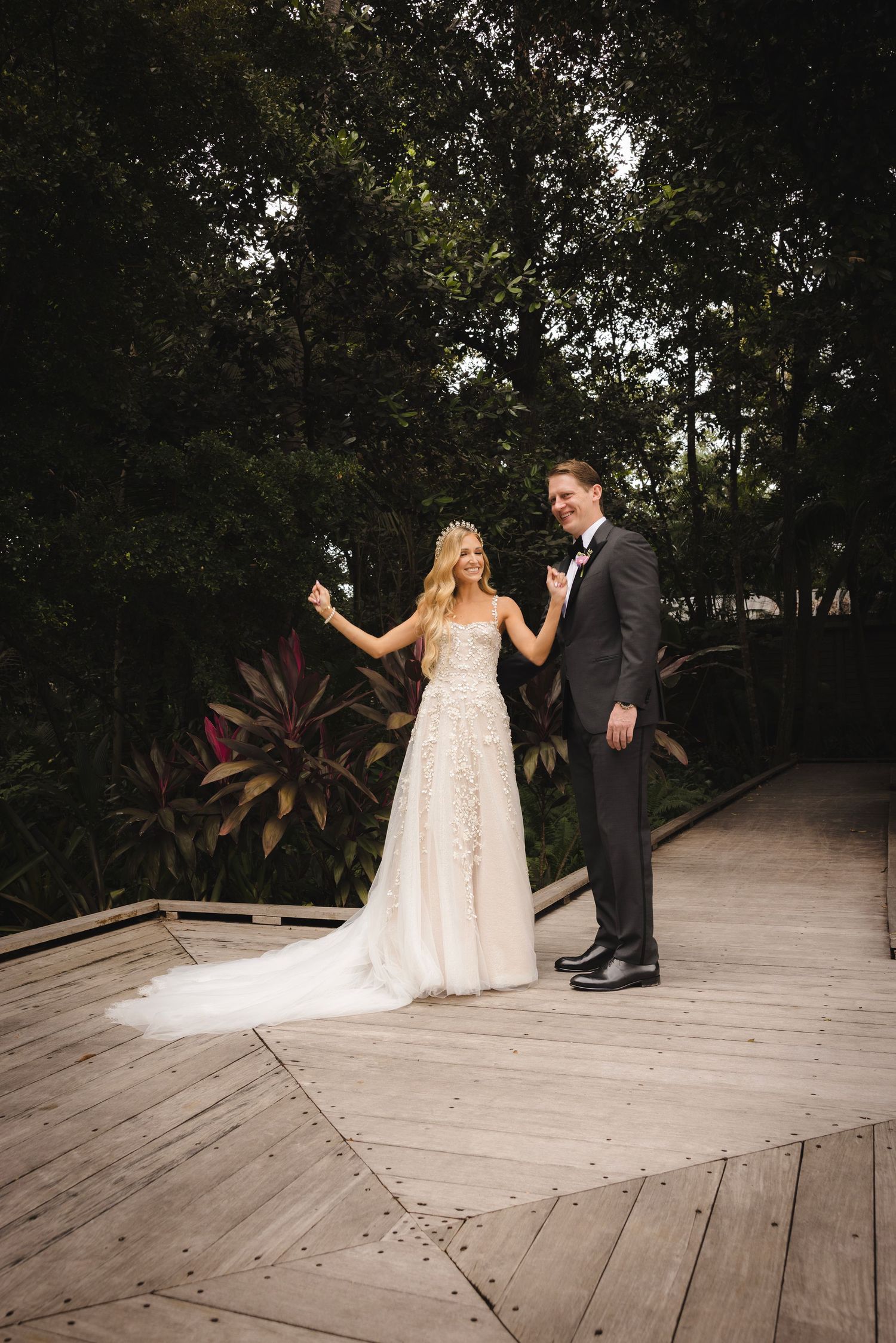 A newlywed couple holds hands on a wooden deck surrounded by lush tropical greenery at their outdoor wedding.