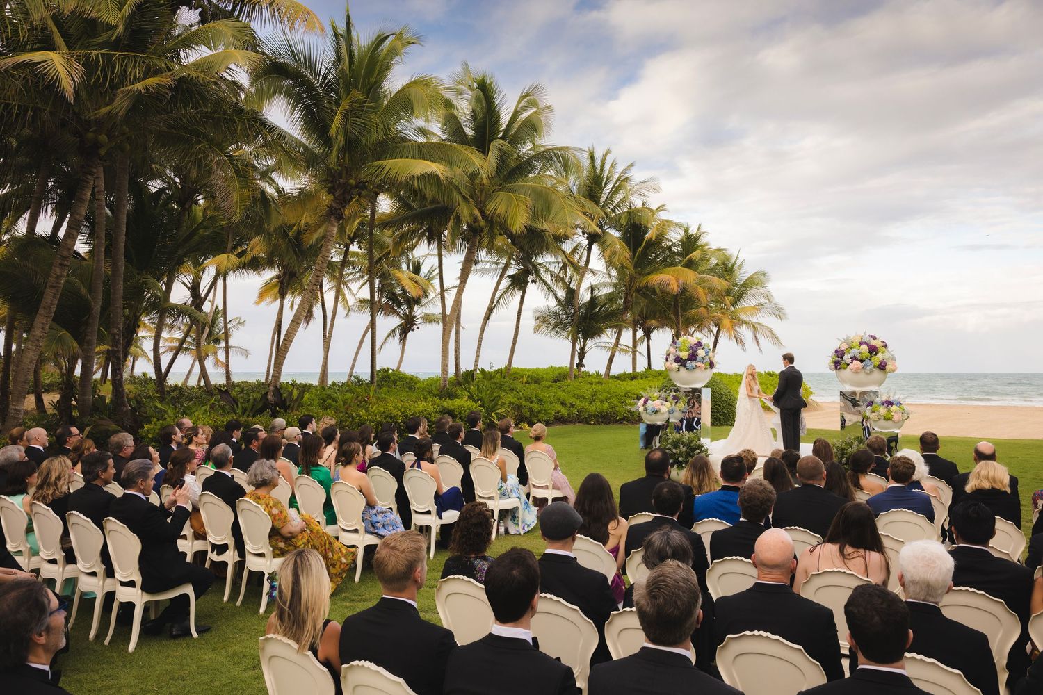 An outdoor beach wedding ceremony in Hawaii with palm trees and white chairs set up facing the ocean under cloudy skies.