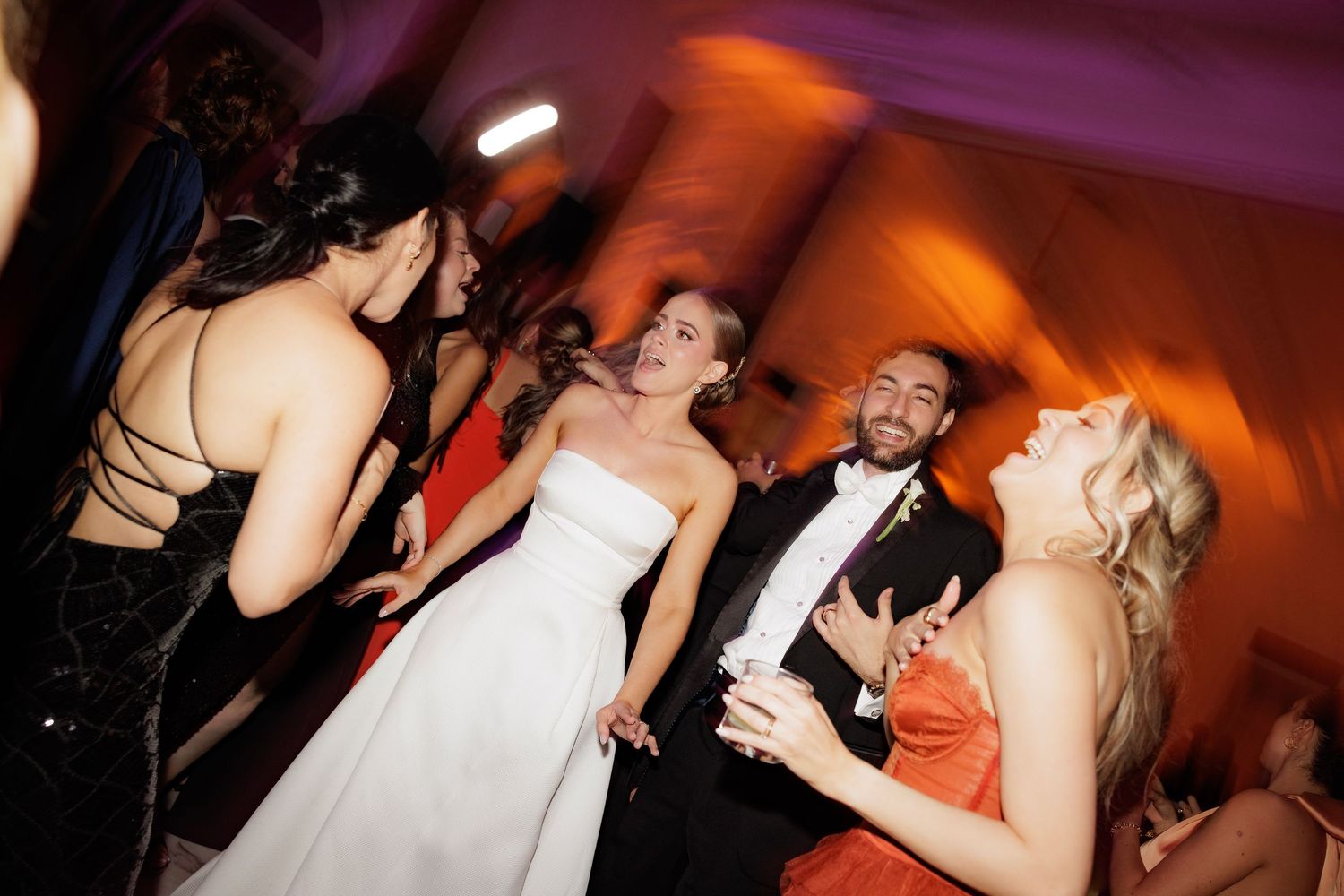 Wedding reception guests dancing and celebrating on a dance floor with orange and purple mood lighting in background.