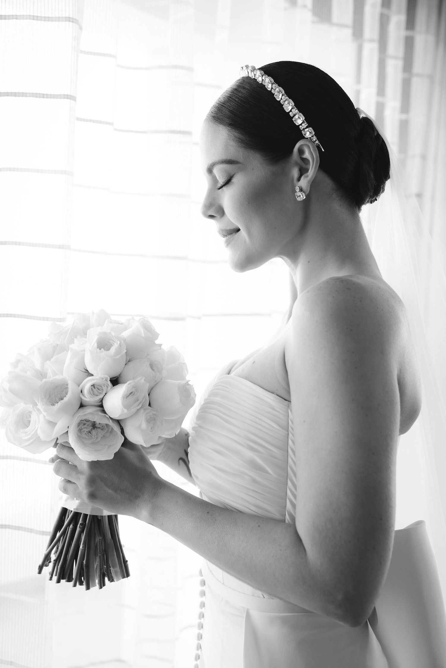 A serene black and white bridal portrait showing a strapless dress and delicate headpiece as she holds a white rose bouquet.