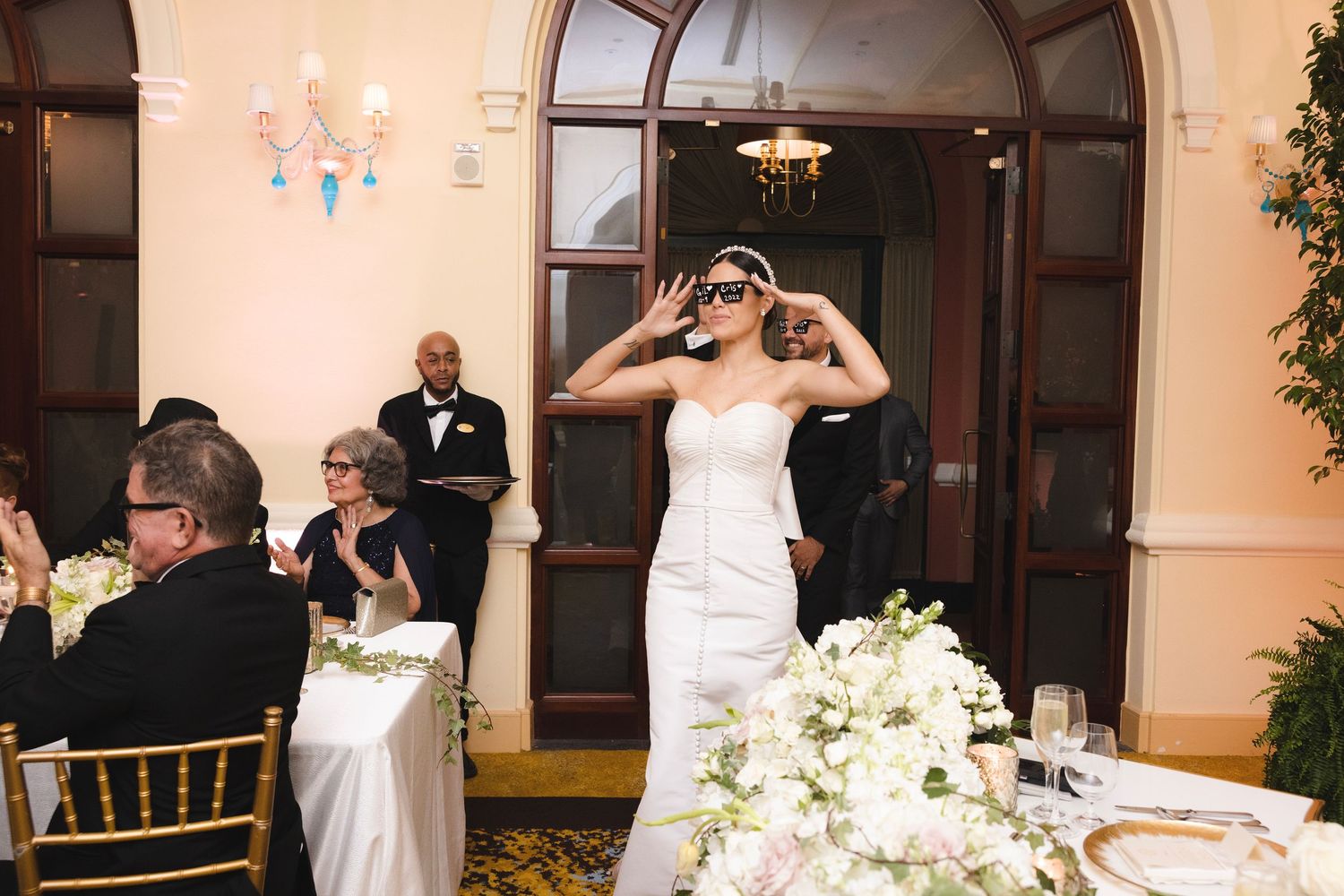 A person in a white wedding dress stands at a reception table decorated with elegant white floral centerpieces.