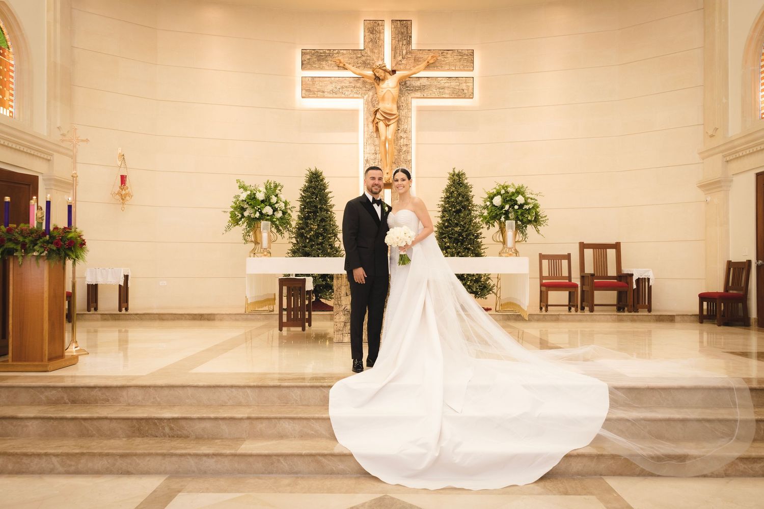 A wedding ceremony in a church sanctuary with white floral arrangements and a large crucifix mounted on the wall.
