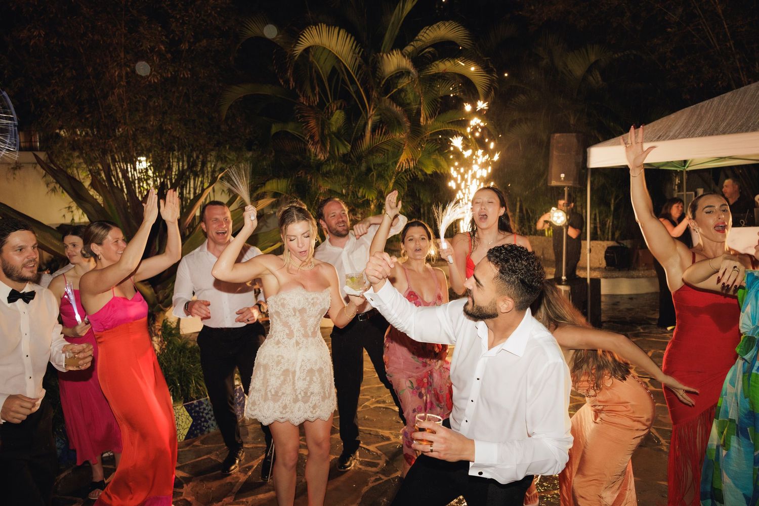 Wedding guests dancing and celebrating at an outdoor tropical reception with palm trees and string lights.