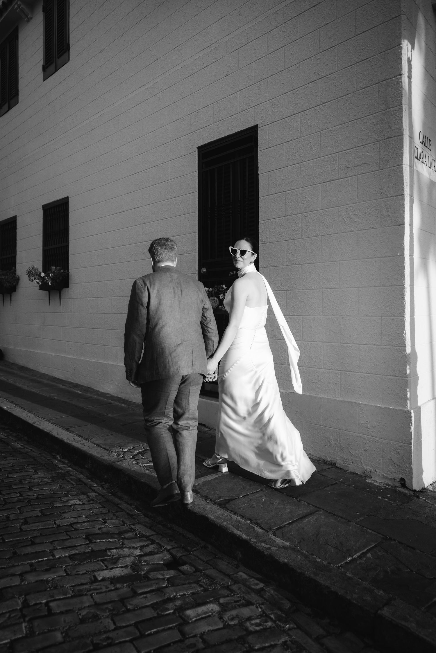 Black and white sequence shows two figures walking down a dim cobblestone corridor beside a white wall.