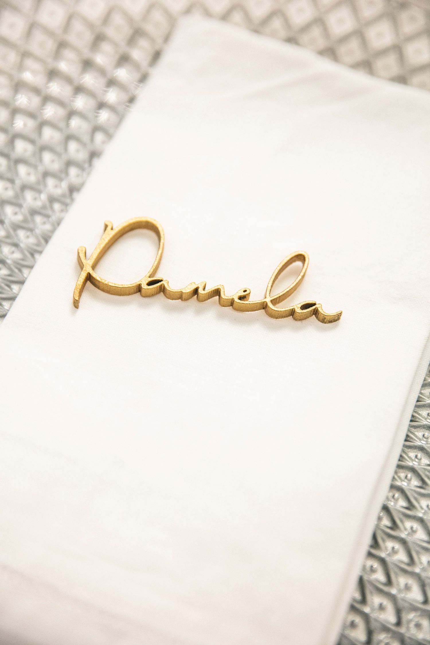 Gold cursive text spelling out 'Pamela' appears elegantly written on white paper with a geometric pattern in background.
