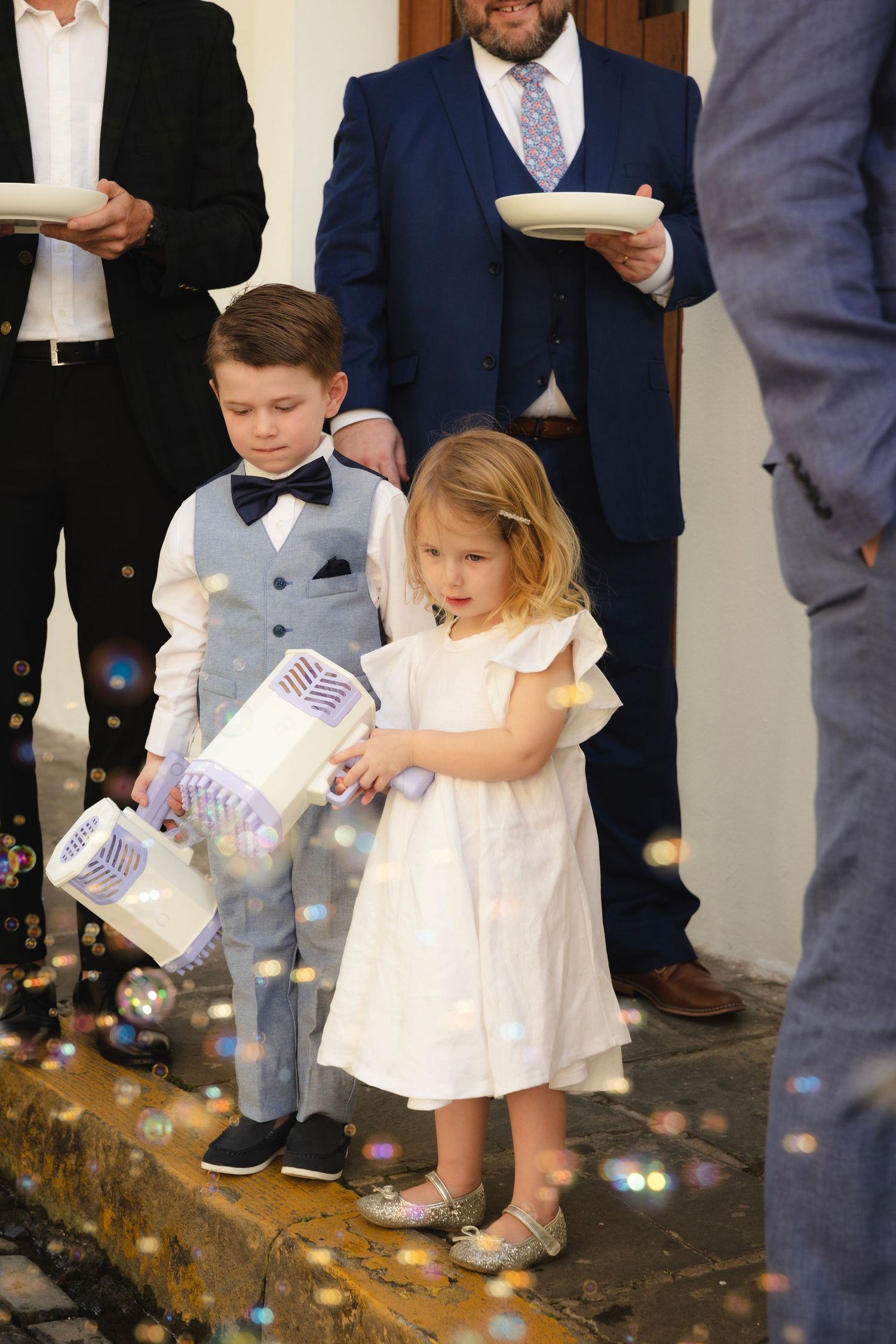 Young children in white outfits spread confetti on the ground during a celebration.