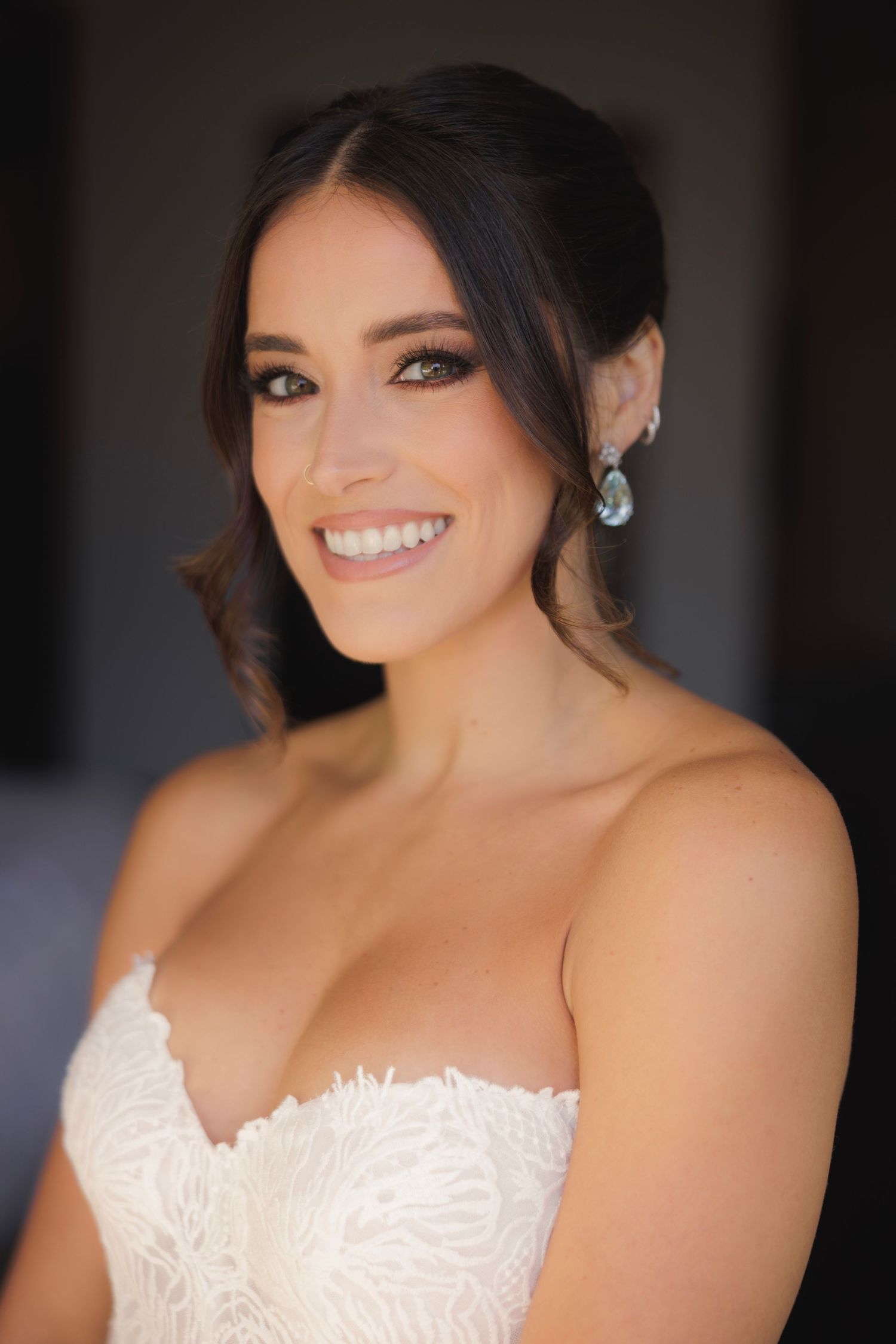 Close-up portrait of a bride in a white lace gown with loose updo hairstyle and sparkling teardrop earrings.