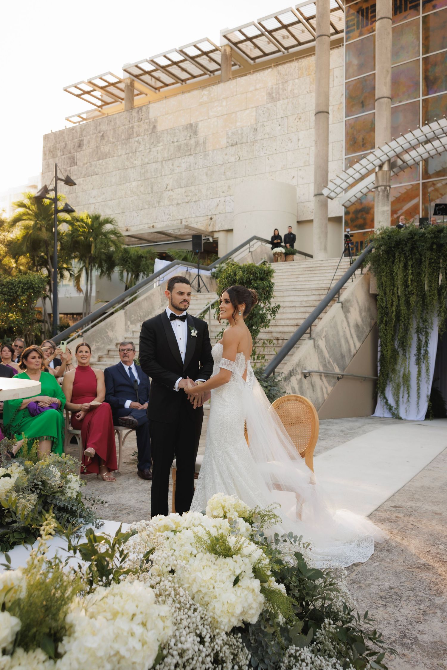 Elegant outdoor wedding ceremony on stone stairs with white floral arrangements and guests in formal attire.