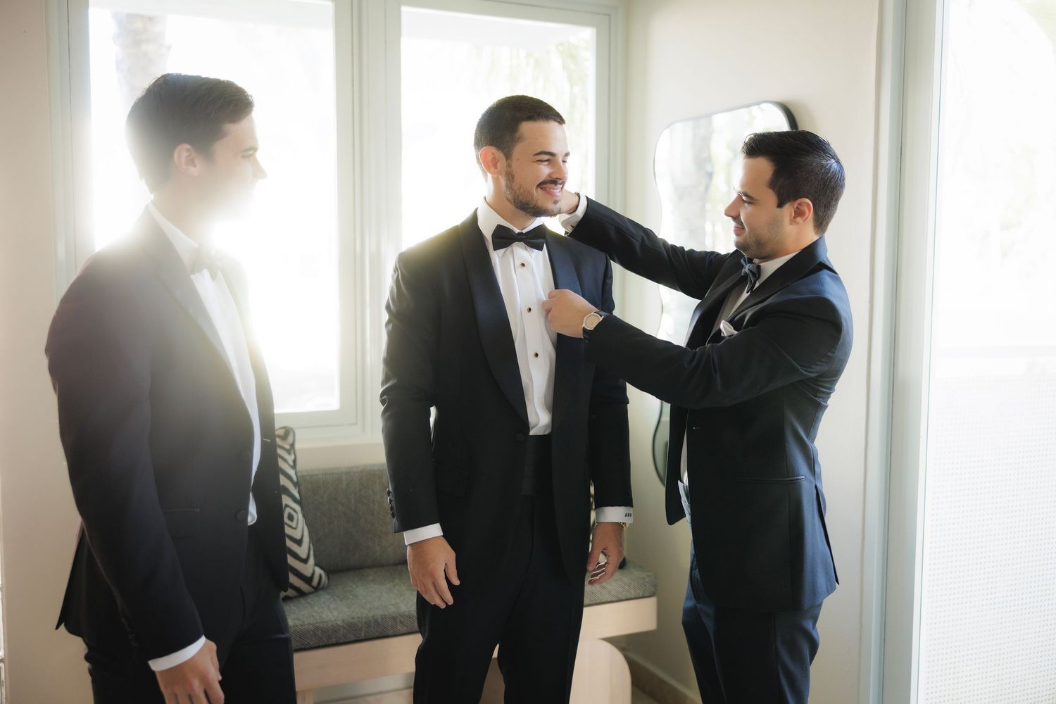 Groomsmen help adjust groom's bowtie in sunlit room before wedding ceremony.
