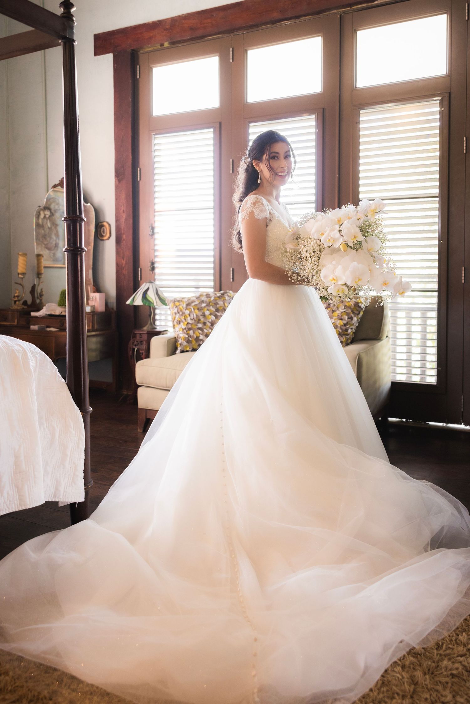 A beautiful flowing white wedding gown with tulle skirt photographed in a sunlit room with windows and wood trim.