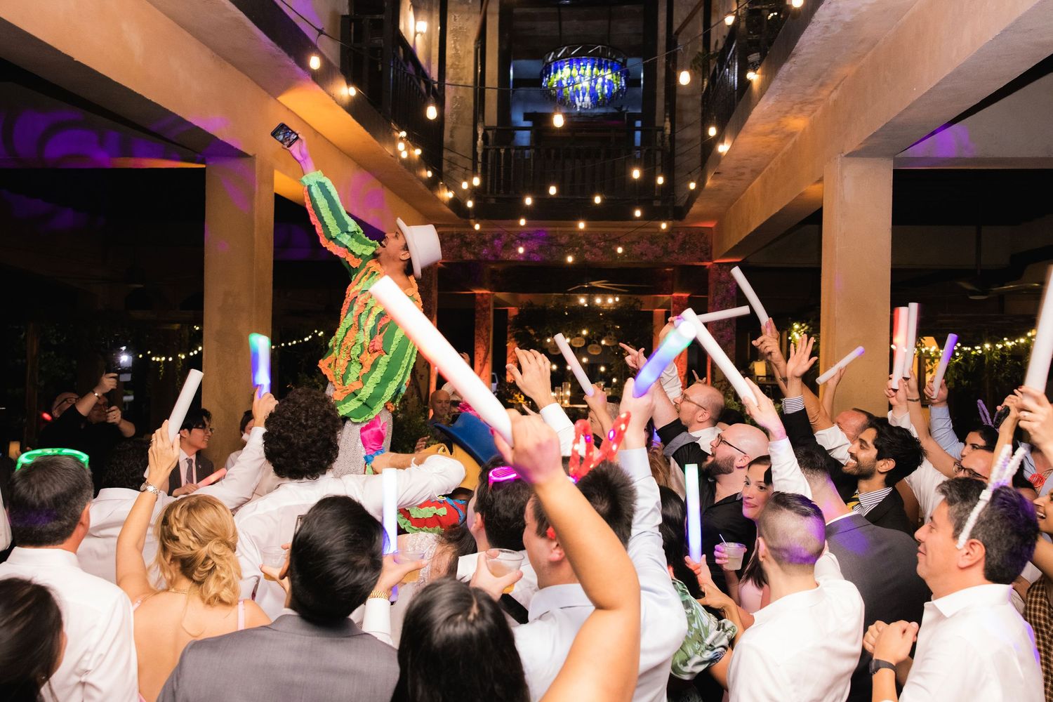 Wedding reception dance party with crowd holding glow sticks and dancing under purple and blue lighting.