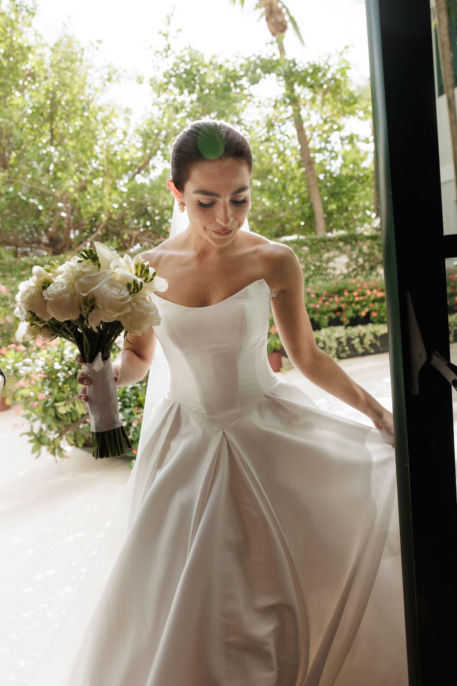 Elegant bride in a classic white ball gown with bow detail holds her dress while standing in natural light.
