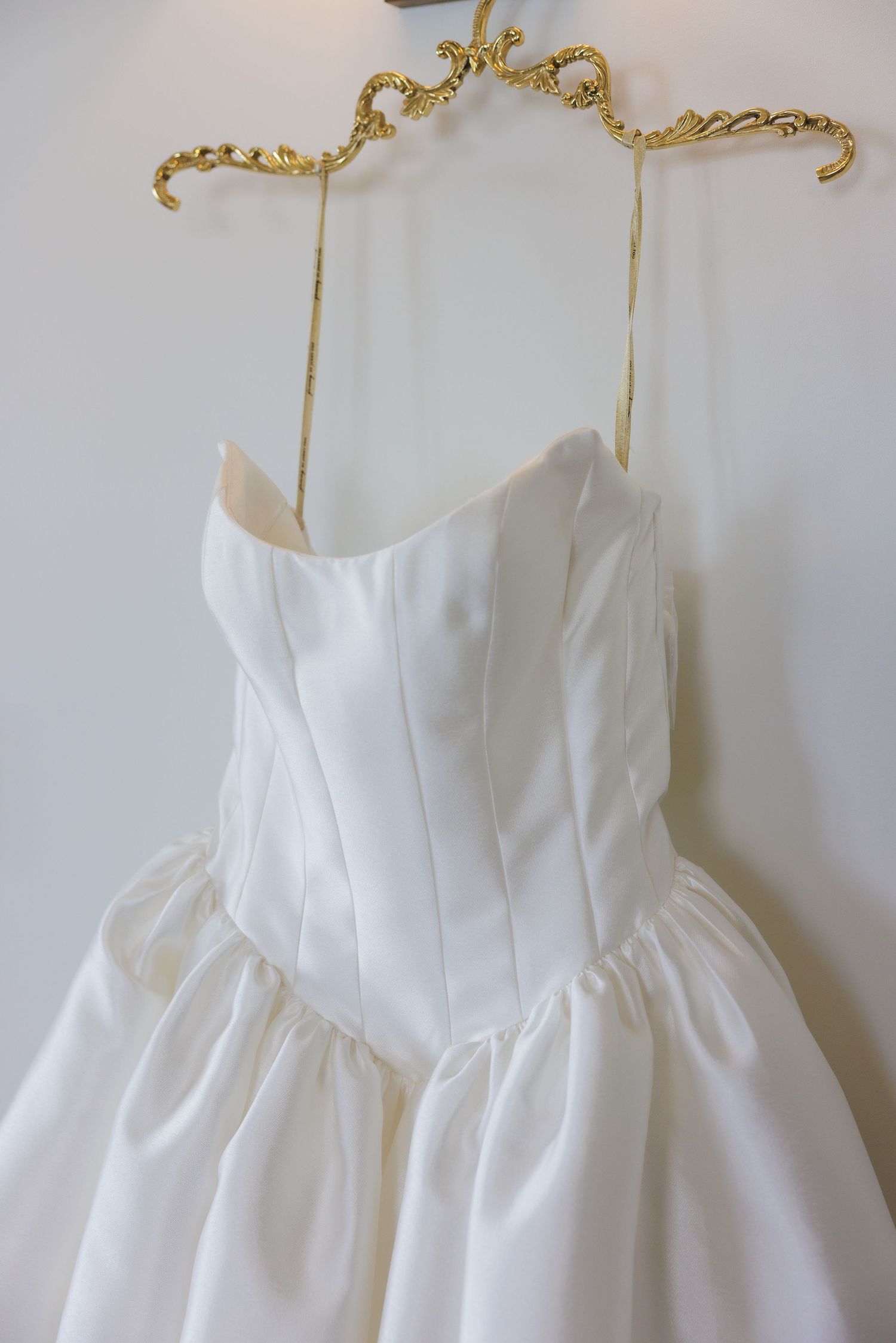 Classic white wedding dress with structured bodice hanging on ornate gold hanger against white wall.