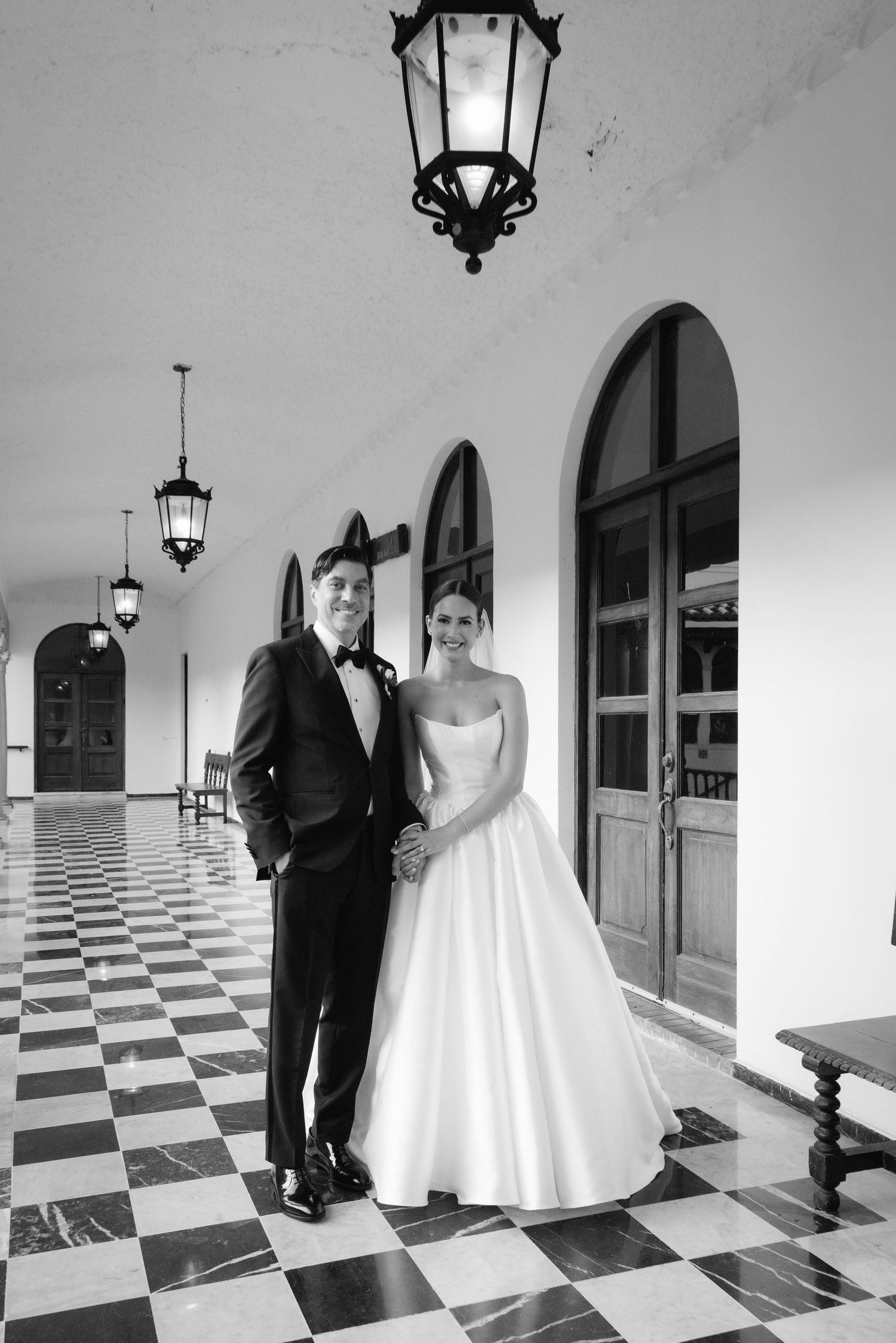 A black and white wedding portrait on a checkered floor with arched windows and hanging lanterns.