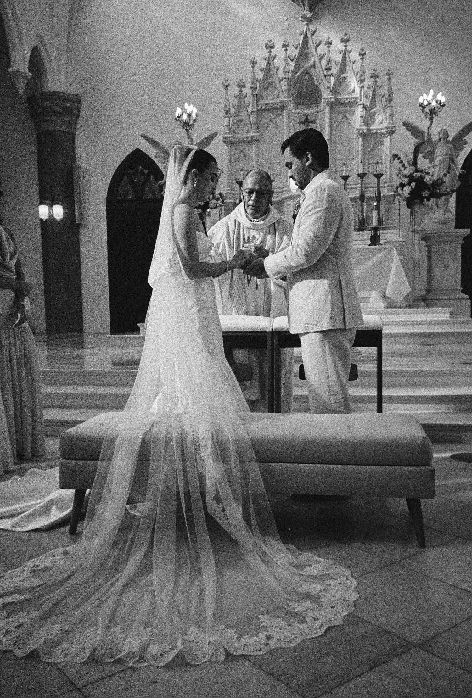 Black and white photo of bride and groom exchanging vows before a priest at an ornate church altar.