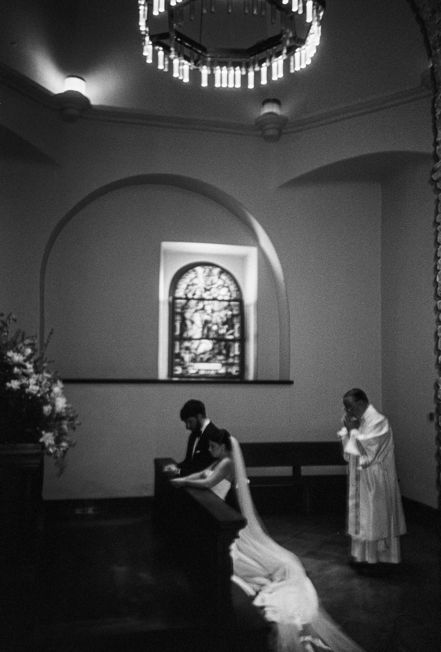 Black and white photo of a couple kneeling at church altar during wedding ceremony with stained glass window backdrop.