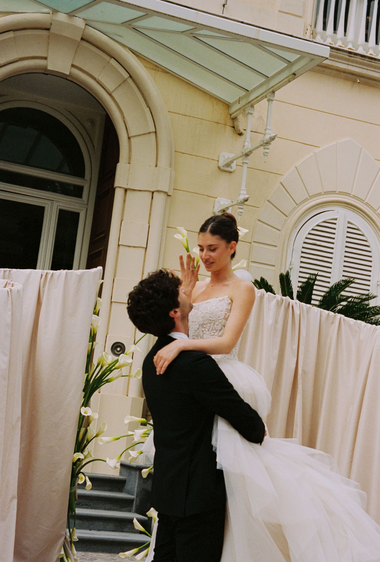 Groom lifts bride in white gown at elegant outdoor wedding venue with arched windows and flowing white drapes.