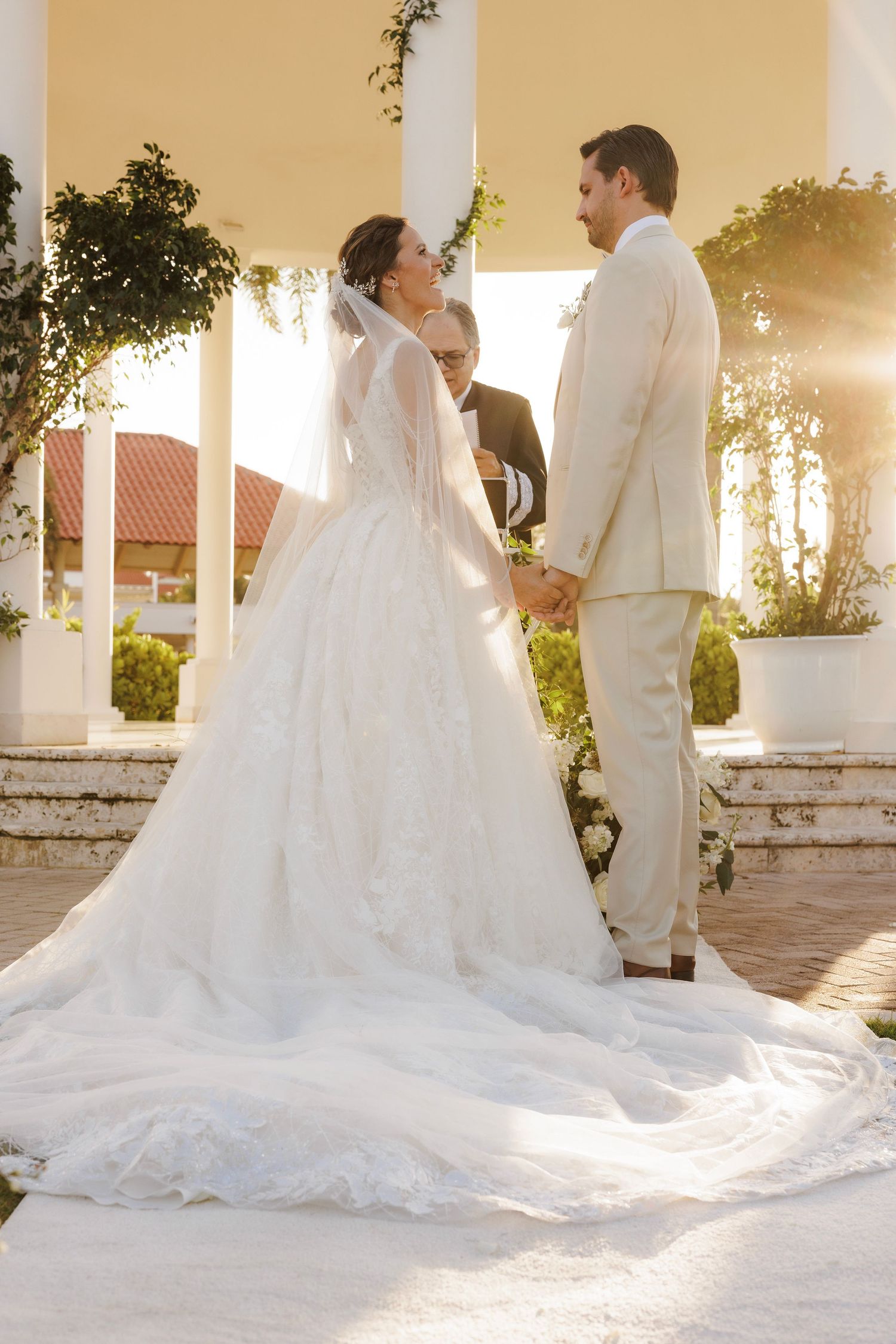 A bride in a flowing white gown and groom in light suit stand together at sunset under elegant white columns.