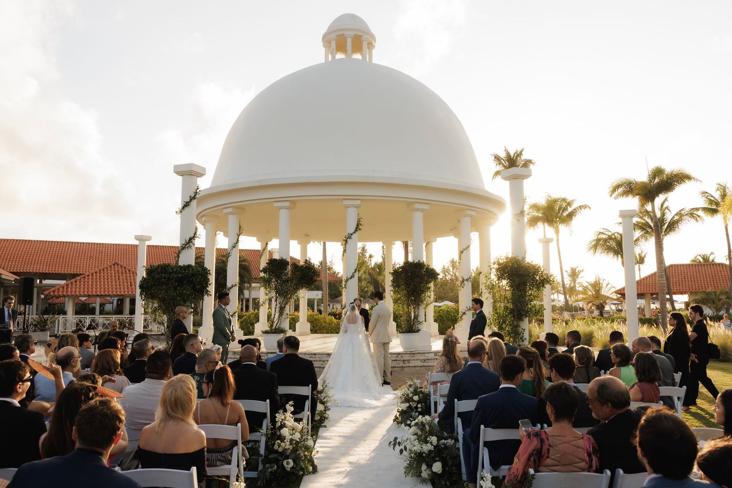 Outdoor beach wedding ceremony under palm trees at sunset with seated guests and a white domed structure.