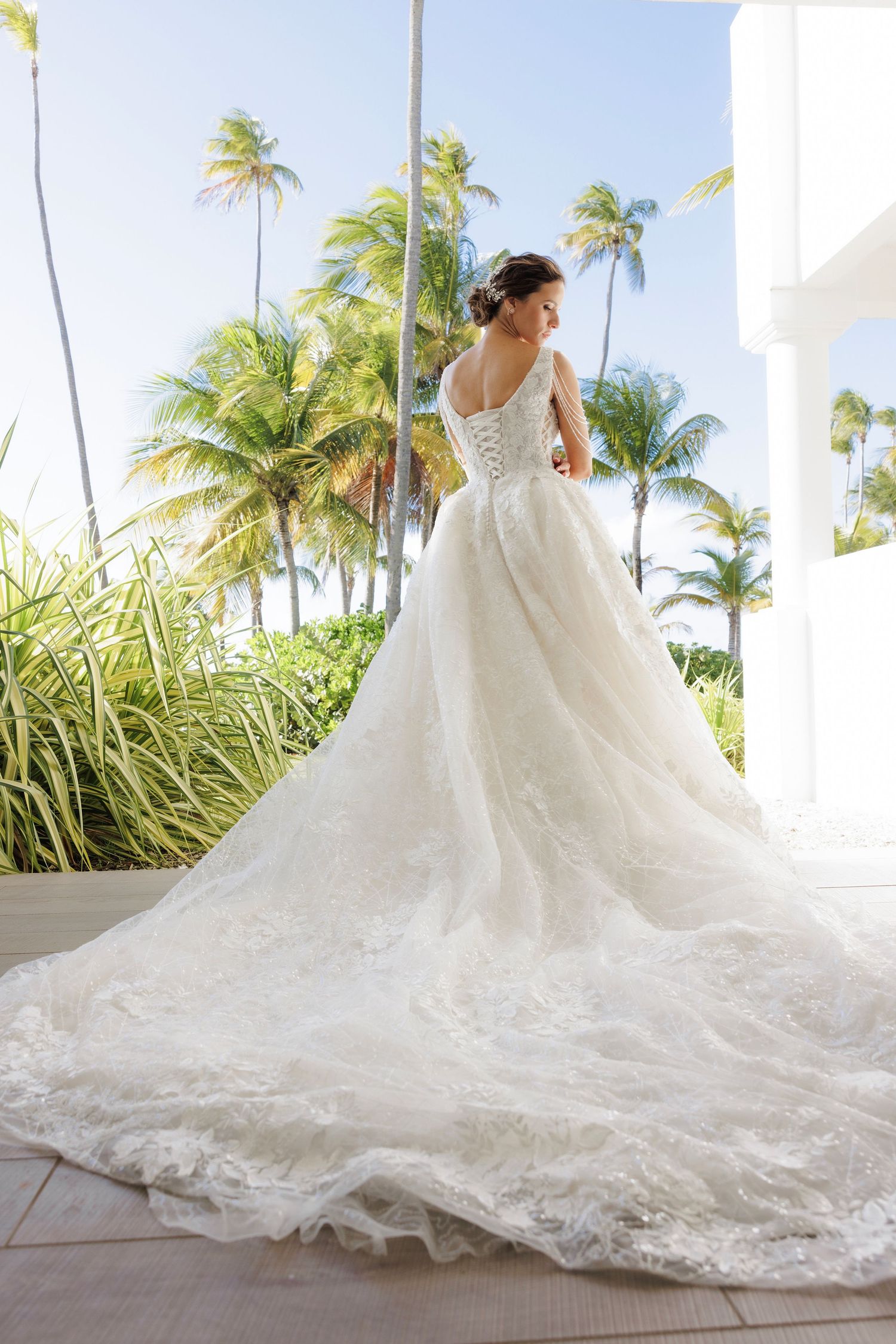 A luxurious white wedding dress with intricate lace detailing and dramatic long train photographed against tropical palm trees.