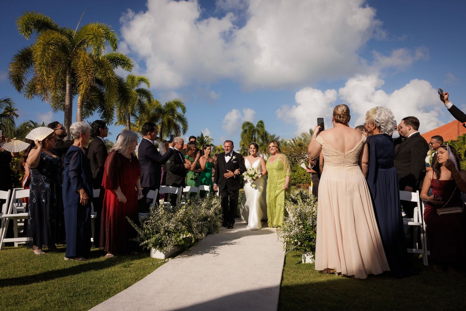 Sequence of wedding ceremony photos showing guests gathered outdoors under blue skies with palm trees and clouds.