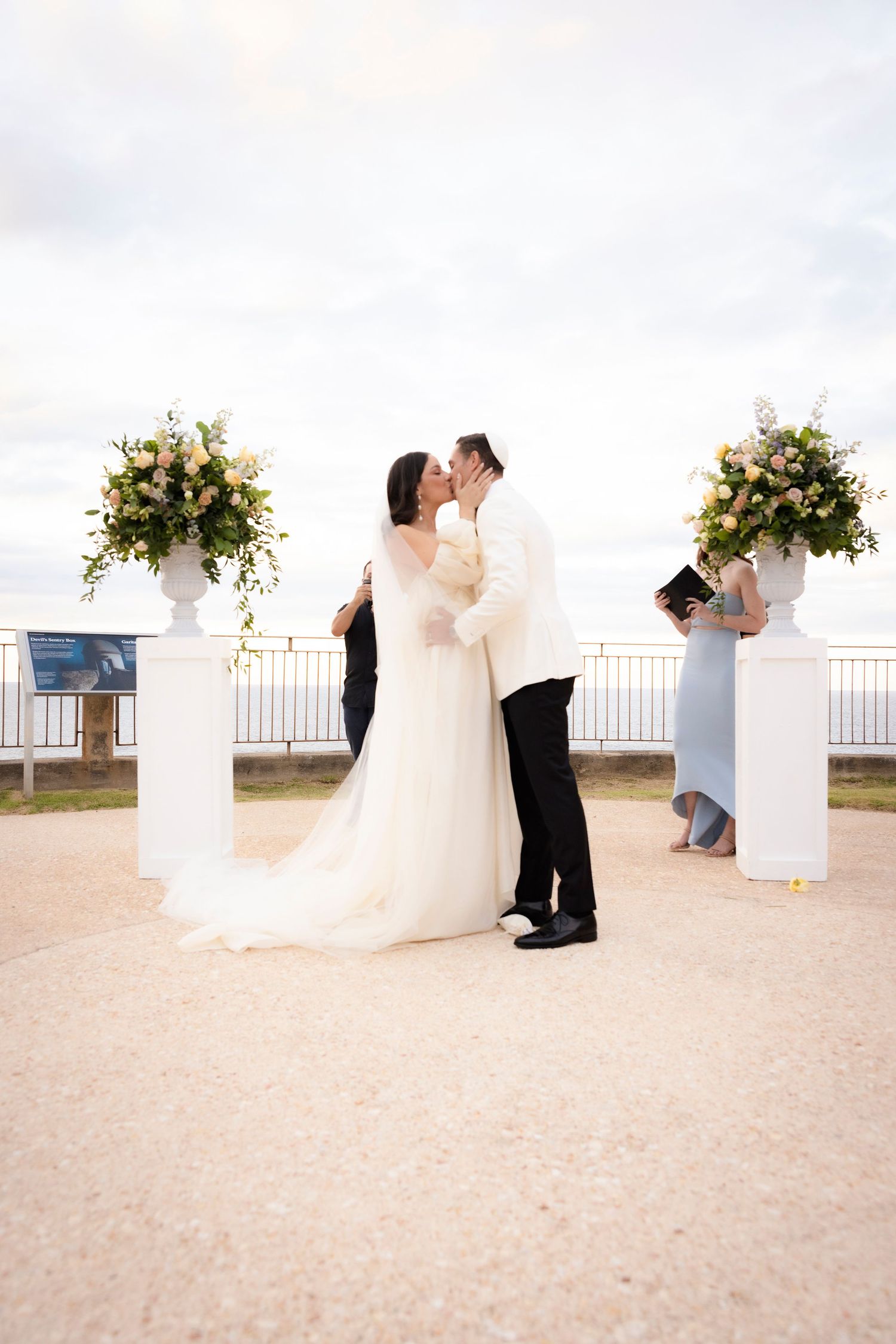 Outdoor wedding ceremony setup with white pedestals and floral arrangements against coastal backdrop.
