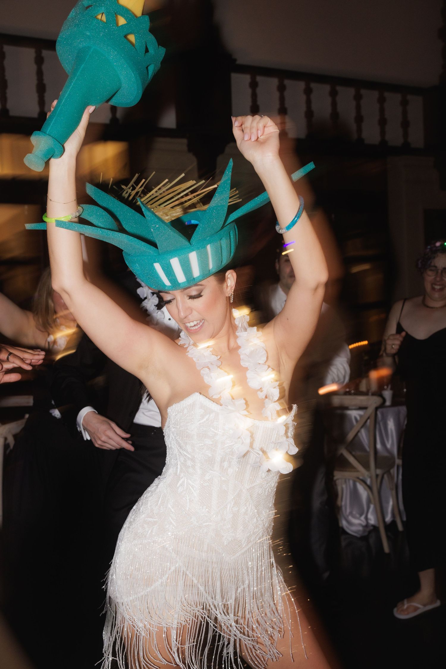 Person in a white fringe dress dances energetically with teal accessories at a dark indoor celebration.