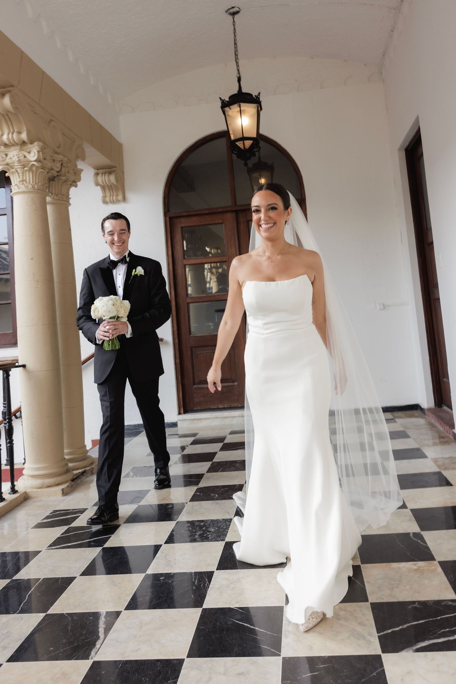 Bride and groom walk through an elegant hallway with black and white checkered floor and white columns.