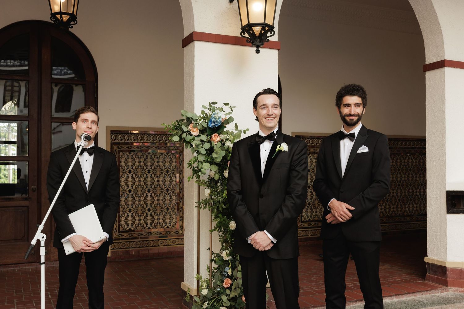 Groomsmen wearing black tuxedos stand together in an elegant archway with decorative lanterns.