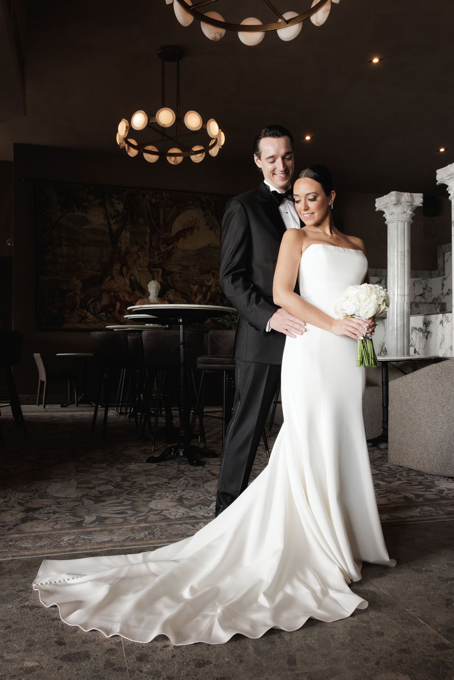 Elegant wedding photo showing a bride in a flowing white gown and groom in black tuxedo embracing in a dimly lit venue.