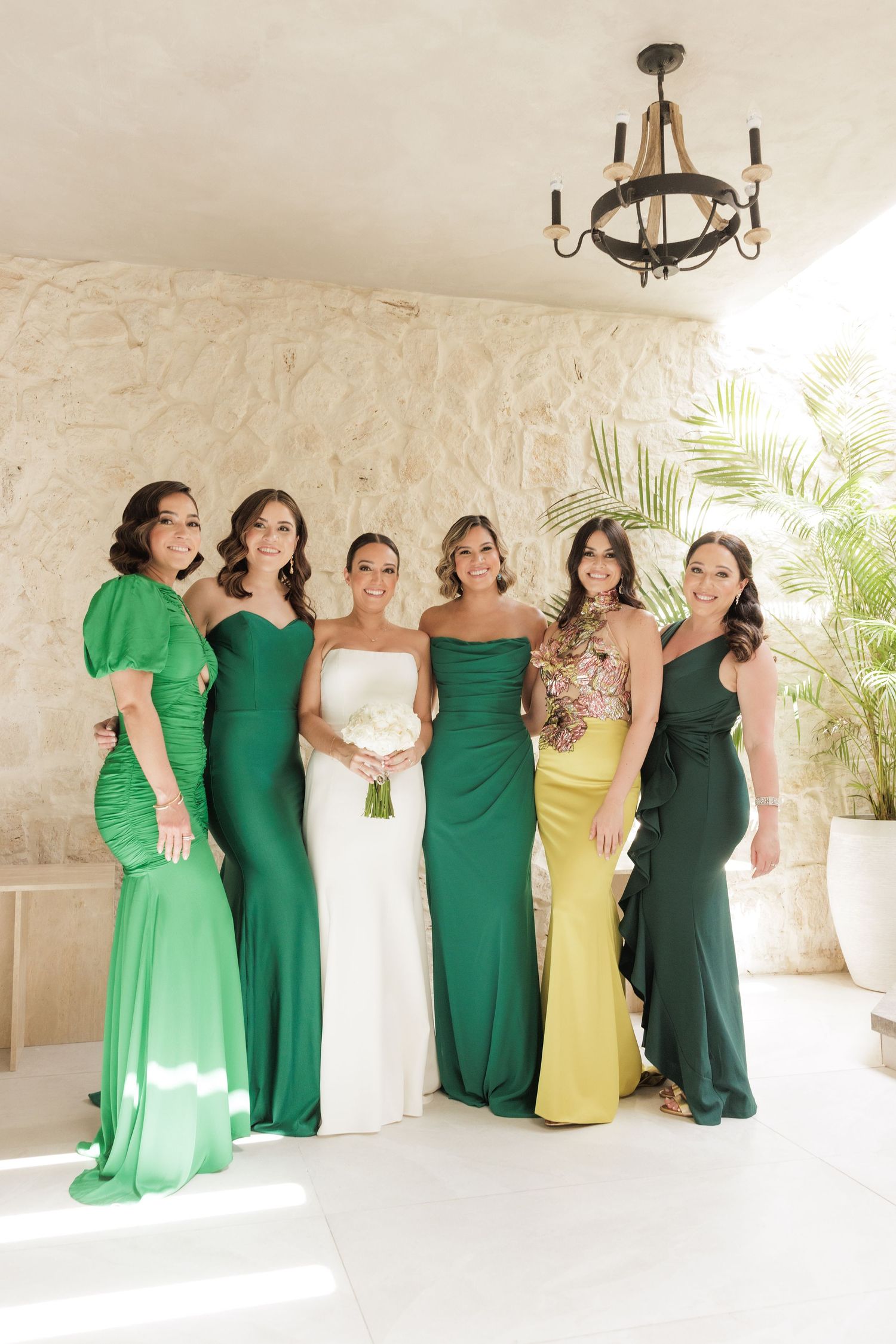 Bridesmaids in emerald green and yellow dresses pose together in an elegant white corridor.