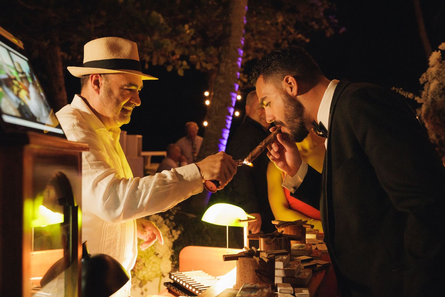 A cigar vendor in a white suit and hat lighting a cigar for a guest at an outdoor evening event.