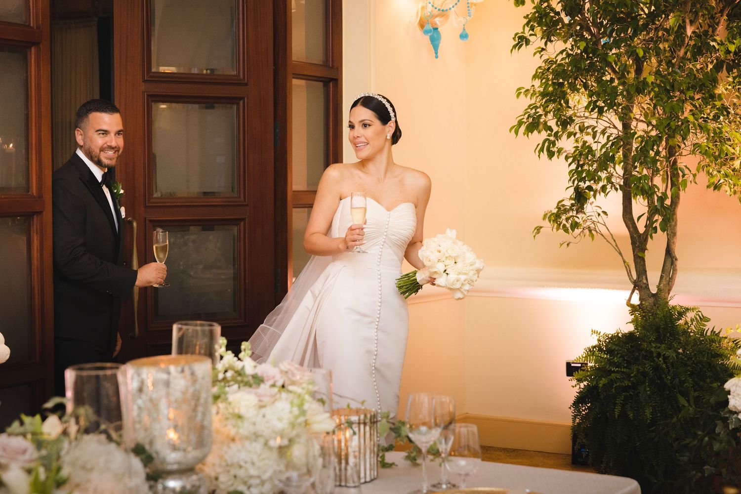 Elegant wedding reception setting with a bride in white gown holding bouquet next to decorative indoor tree.